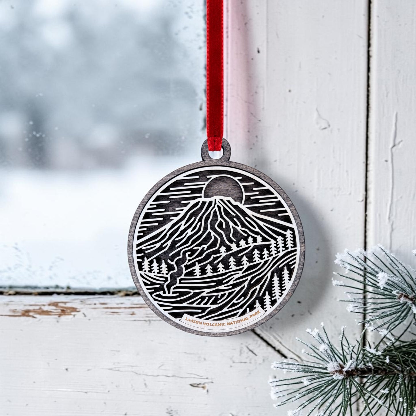 The Lassen Volcanic National Park Line Art Christmas Ornament by Mirabilia Boutique, featuring an oval wooden design with black and white artwork inspired by Lassen Peak, hangs from a red ribbon in a frosty window; a snowy evergreen sprig accentuates the corner while a winter scene unfolds outside.