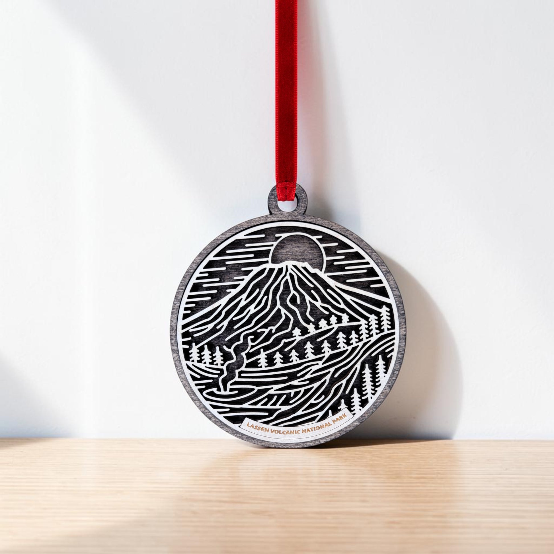 The Lassen Volcanic National Park Line Art Christmas Ornament by Mirabilia Boutique features a round wooden design with black and white line art depicting sun, trees, and a mountain scene. Hanging from a red ribbon, it showcases "Lassen Volcanic National Park" at the bottom to celebrate the beauty of Lassen Peak.
