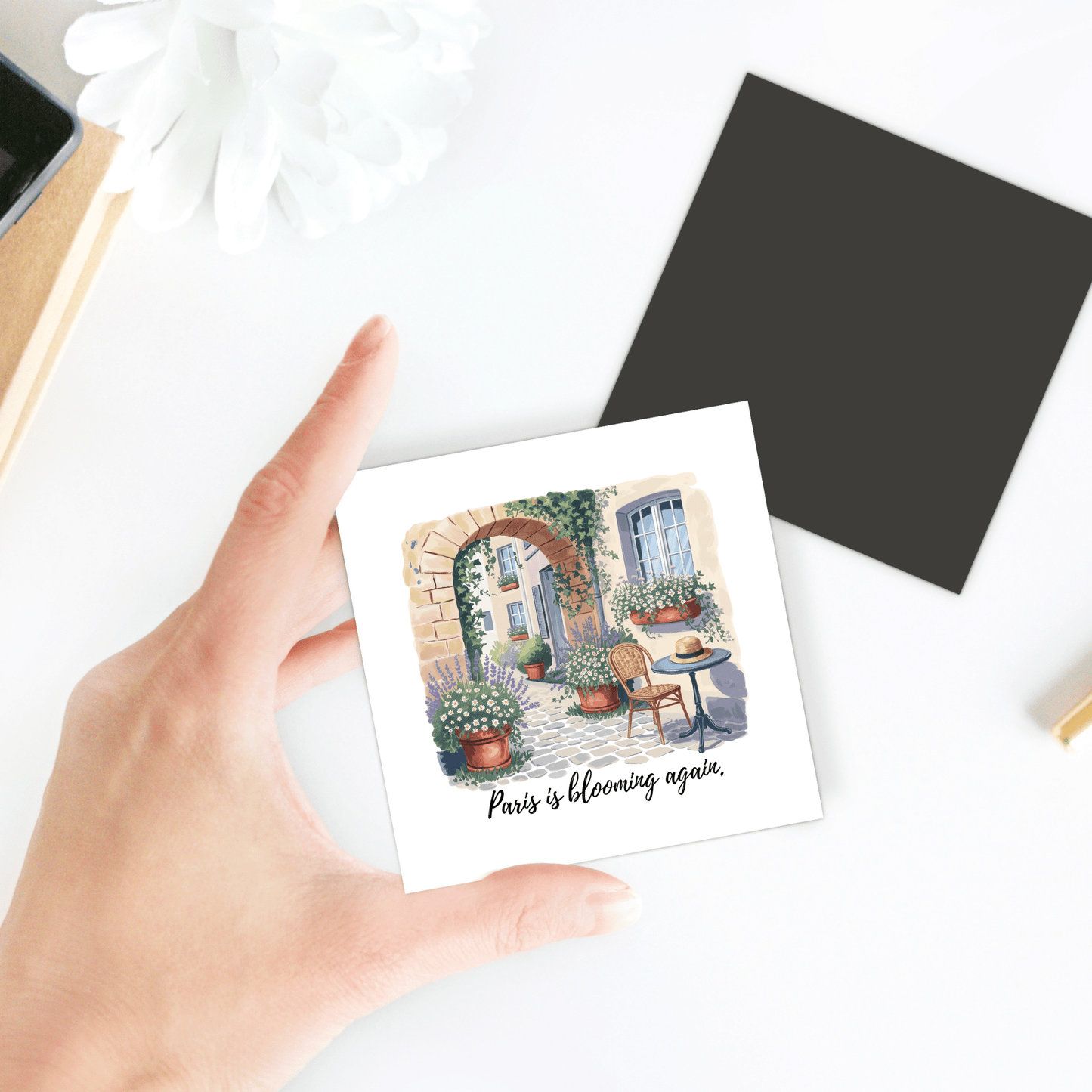 A hand holds Mirabilia Boutique’s “Paris Is Blooming Again – Spring Courtyard Magnet,” featuring potted plants, a chair, and table with the phrase “Paris is blooming again”—a charming spring kitchen décor piece.