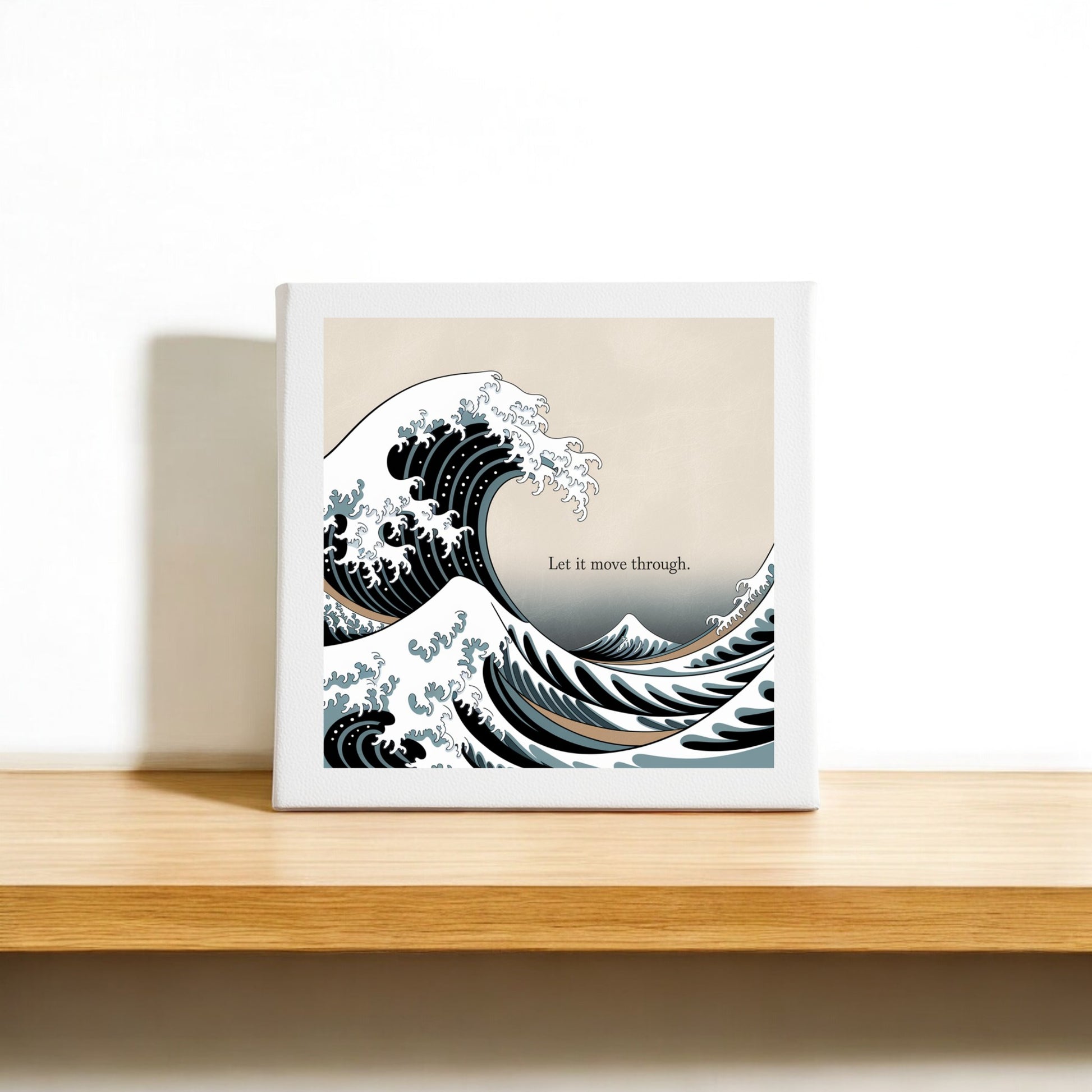 The Let It Move Through Canvas – Bold Ocean Calm Art by Mirabilia Boutique sits on a wooden shelf, featuring the phrase “Let it move through.” Inspired by Japanese ocean art, its plain, minimal background creates a calming effect and evokes emotional resilience.