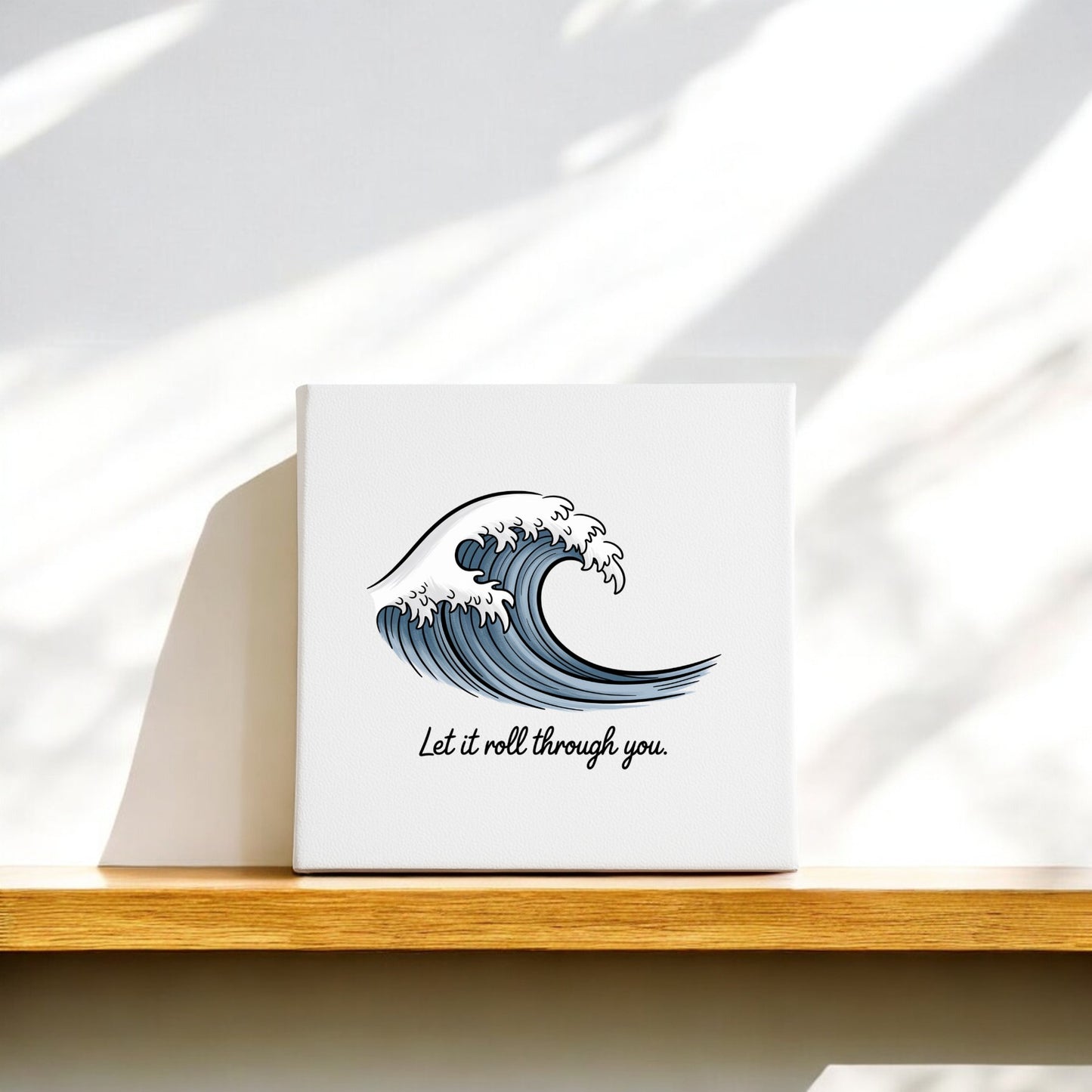 The Let It Roll Through You Canvas – Ocean-Inspired Calm by Mirabilia Boutique features a stylized blue wave and uplifting text, making it soothing meditation decor, ideal for displaying on a shelf with gentle sunlight and shadows.