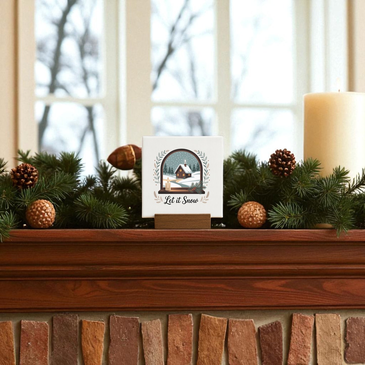 A festive mantel features Mirabilia Boutique’s "Let It Snow Mini Sign"—a 4x4 winter canvas with wood base—amid pine branches, pinecones, and a candle, framed by a window overlooking bare trees. Perfect winter decor!.