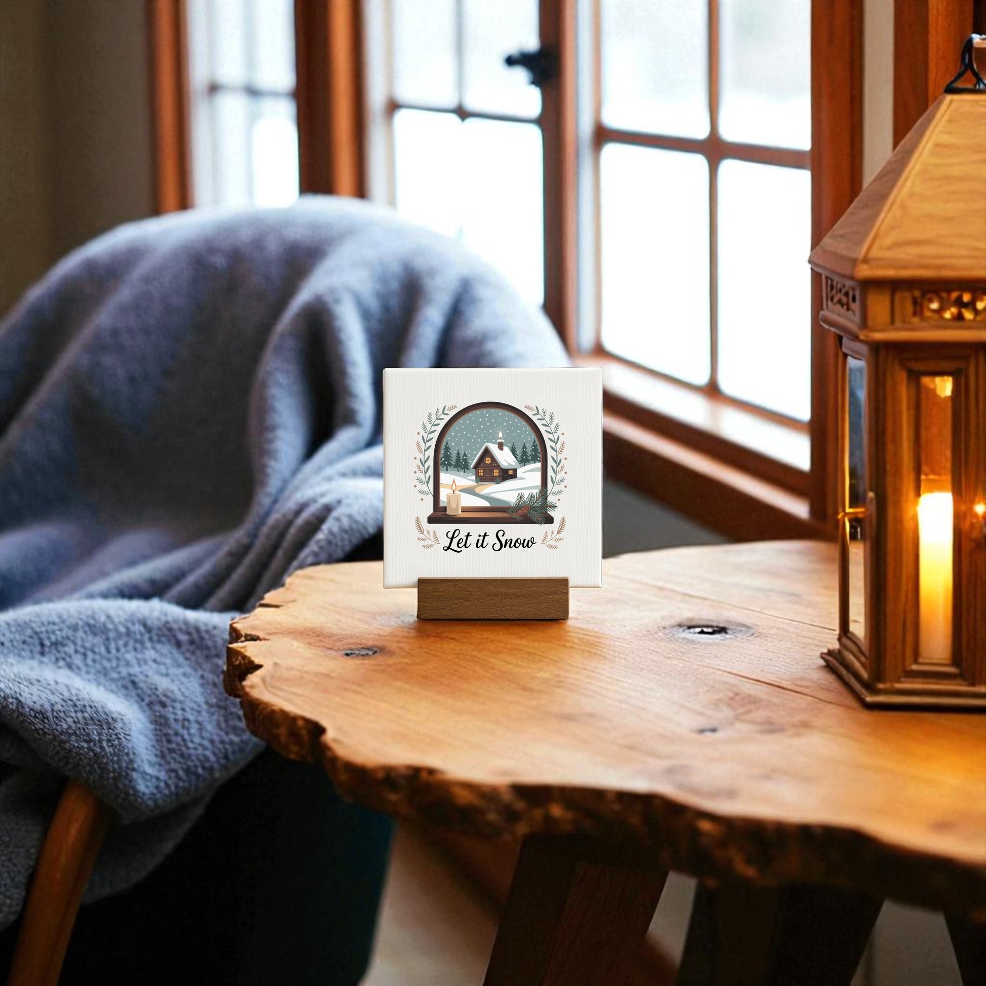 A cozy living room showcases the Mirabilia Boutique Let It Snow Mini Sign, a 4x4 winter canvas with wood base—ideal as charming holiday decor or a gift—set on a wooden table by a blue-draped chair under sunlit windows.