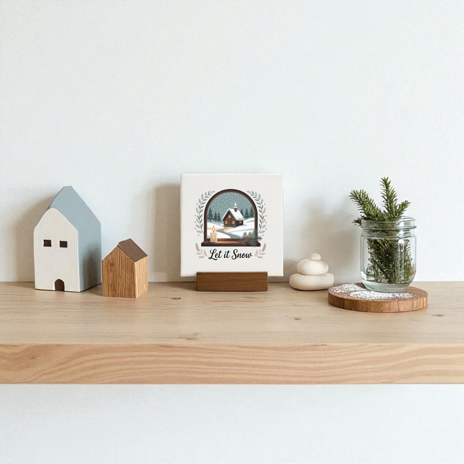 A wooden shelf features two decorative houses, stacked stones, a jar with evergreens, a doily-topped wood coaster, and the Mirabilia Boutique Let It Snow Mini Sign—an ideal winter accent or charming holiday gift.
