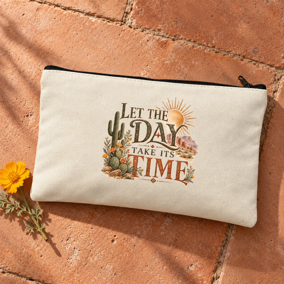 The Let the Day Take Its Time Makeup Bag | Sage & Stone by Mirabilia Boutique features a desert cactus design, sun, wildflowers, and hills. Shown on terracotta tiles with a yellow flower beside it.