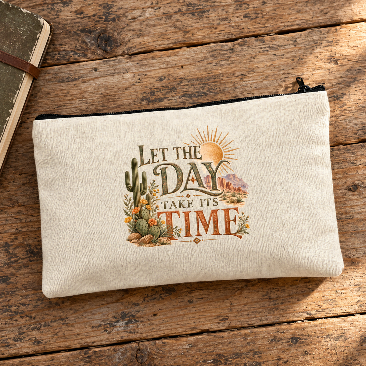 The Let the Day Take Its Time Makeup Bag | Sage & Stone by Mirabilia Boutique in beige, with desert plants and sunrise design, sits on a rustic wooden surface next to a notebook.