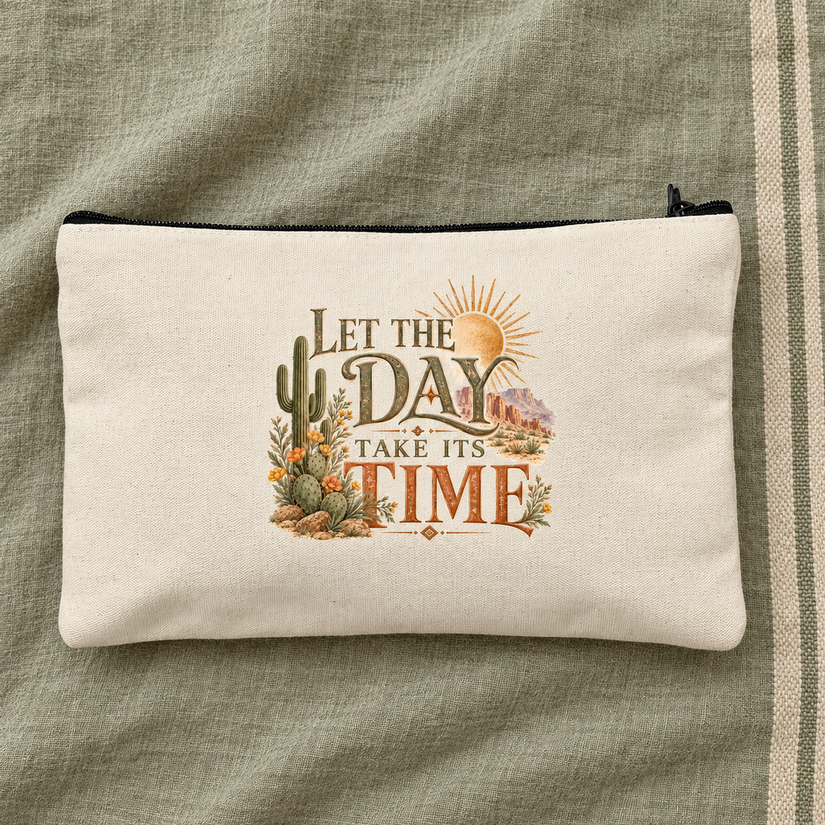 The “Let the Day Take Its Time Makeup Bag | Sage & Stone” by Mirabilia Boutique features a serene desert scene with sun, cactus, and flowers on beige fabric, printed with “Let the day take its time.”.