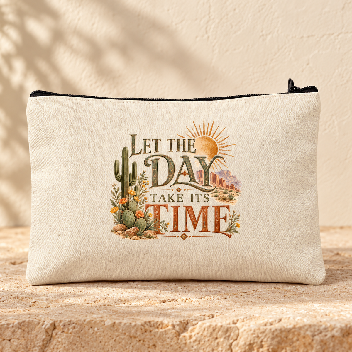 The Let the Day Take Its Time Makeup Bag | Sage & Stone by Mirabilia Boutique features charming cactus art and an inspiring quote, styled as a decorative zipper pouch on a textured stone surface.