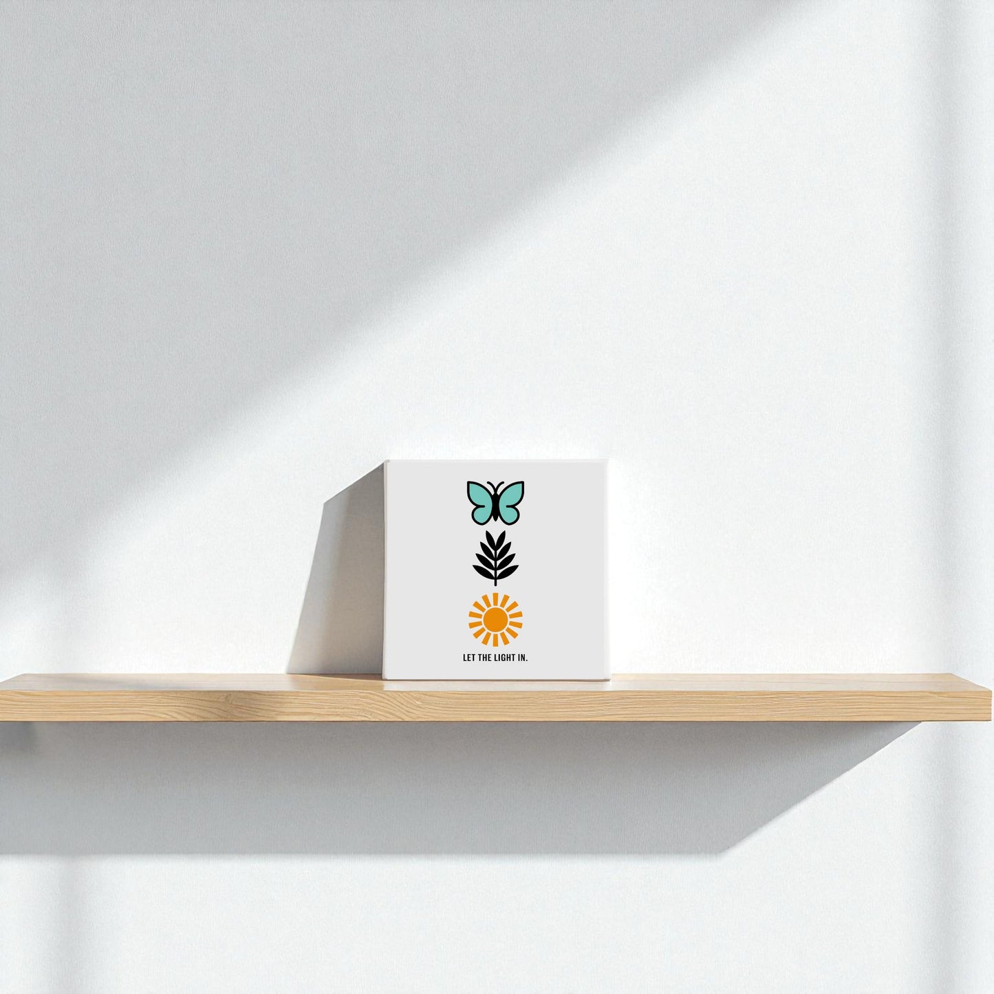 The Let the Light In Canvas – Butterfly Sun Wall Art by Mirabilia Boutique sits on a light wooden shelf against a white wall, its minimalist butterfly, leaf, and sun design brightened by sunlight to create an inspiring decor accent.