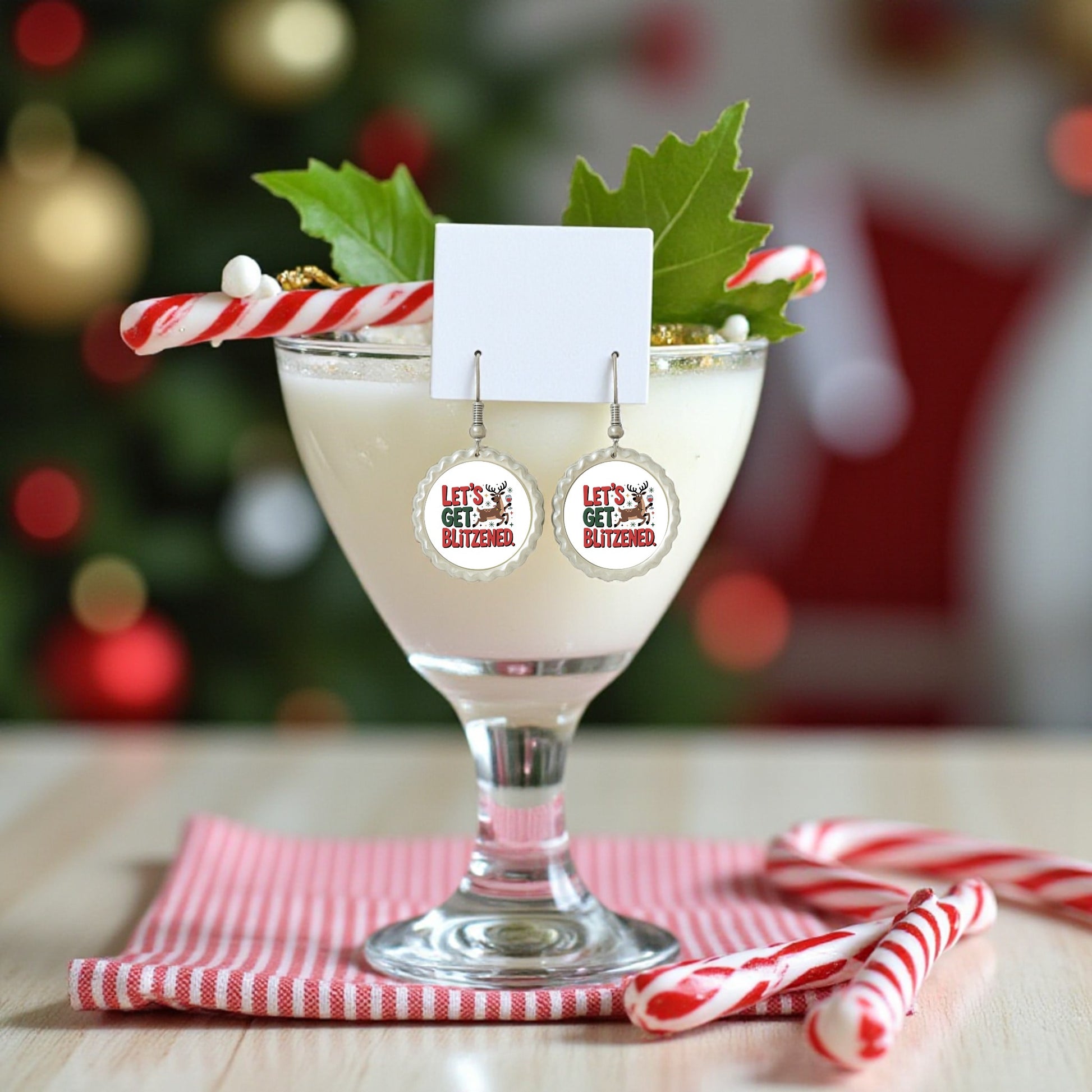 A festive holiday drink with a candy cane, holly, and gold accents sits beside red-and-white candy canes and a striped napkin. In front are Mirabilia Boutique’s "Let's Get Blitzened" funny Christmas earrings.
