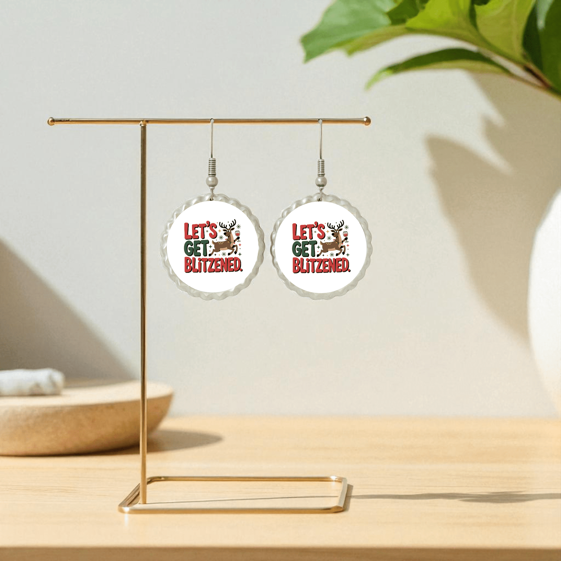 Mirabilia Boutique's Let's Get Blitzened | Funny Christmas Earrings feature reindeer heads and festive text, shown on a gold stand atop a wooden surface with a green plant in the background—perfect for your next holiday party.