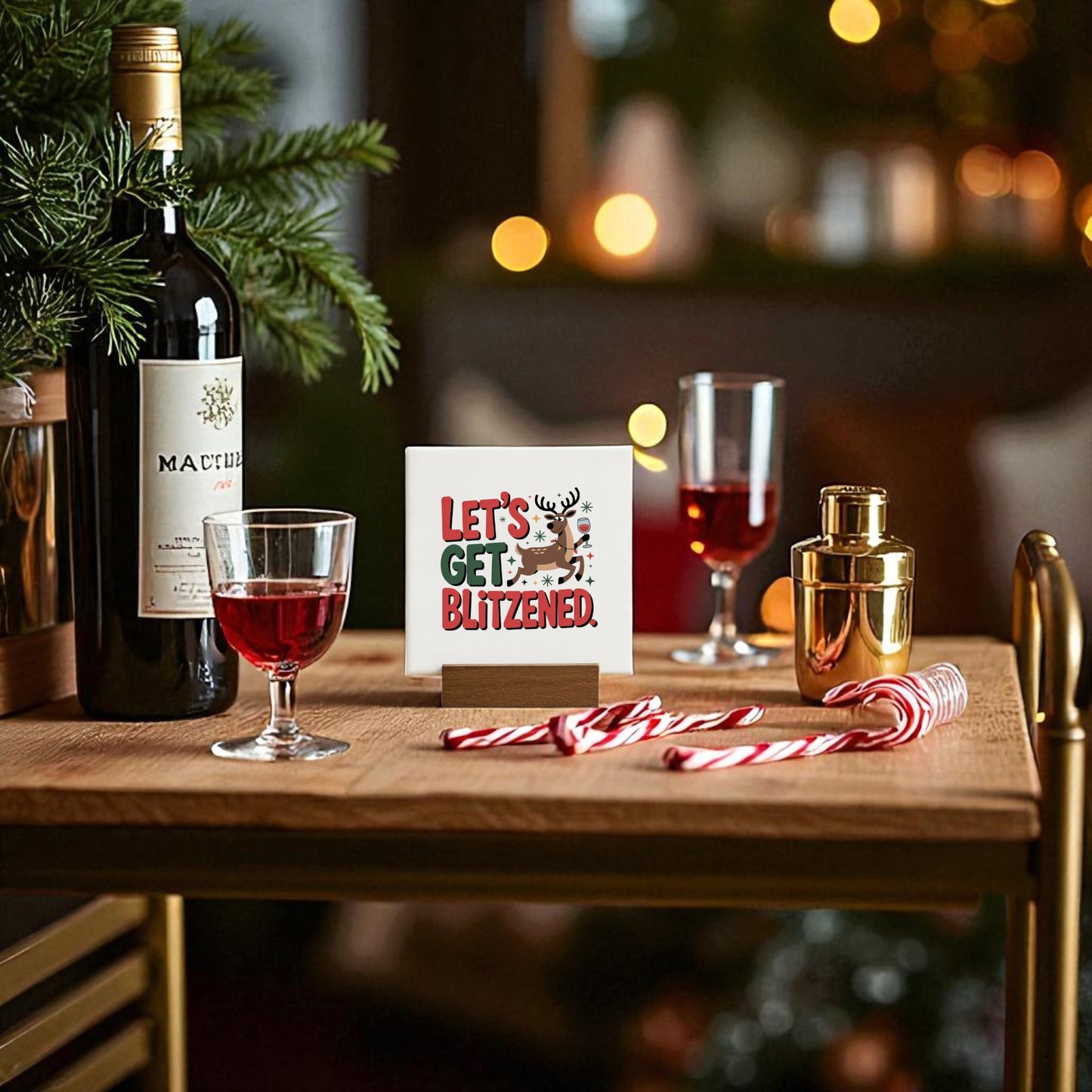 A festive table features a bottle of red wine, two glasses, a gold cocktail shaker, candy canes, and Mirabilia Boutique's "Let's Get Blitzened" Mini Christmas Sign. Holiday lights and greenery create cozy Christmas decor for this inviting scene.