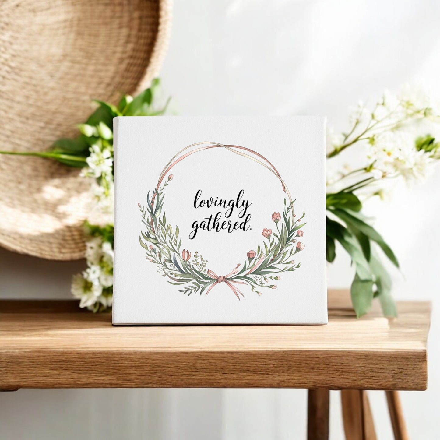The Mirabilia Boutique Lovingly Gathered Canvas – 8x8 Botanical Wreath Wall Art, hand-painted with a floral wreath and “lovingly gathered,” sits on a wooden table, accented by white flowers and a woven straw hat in the background.