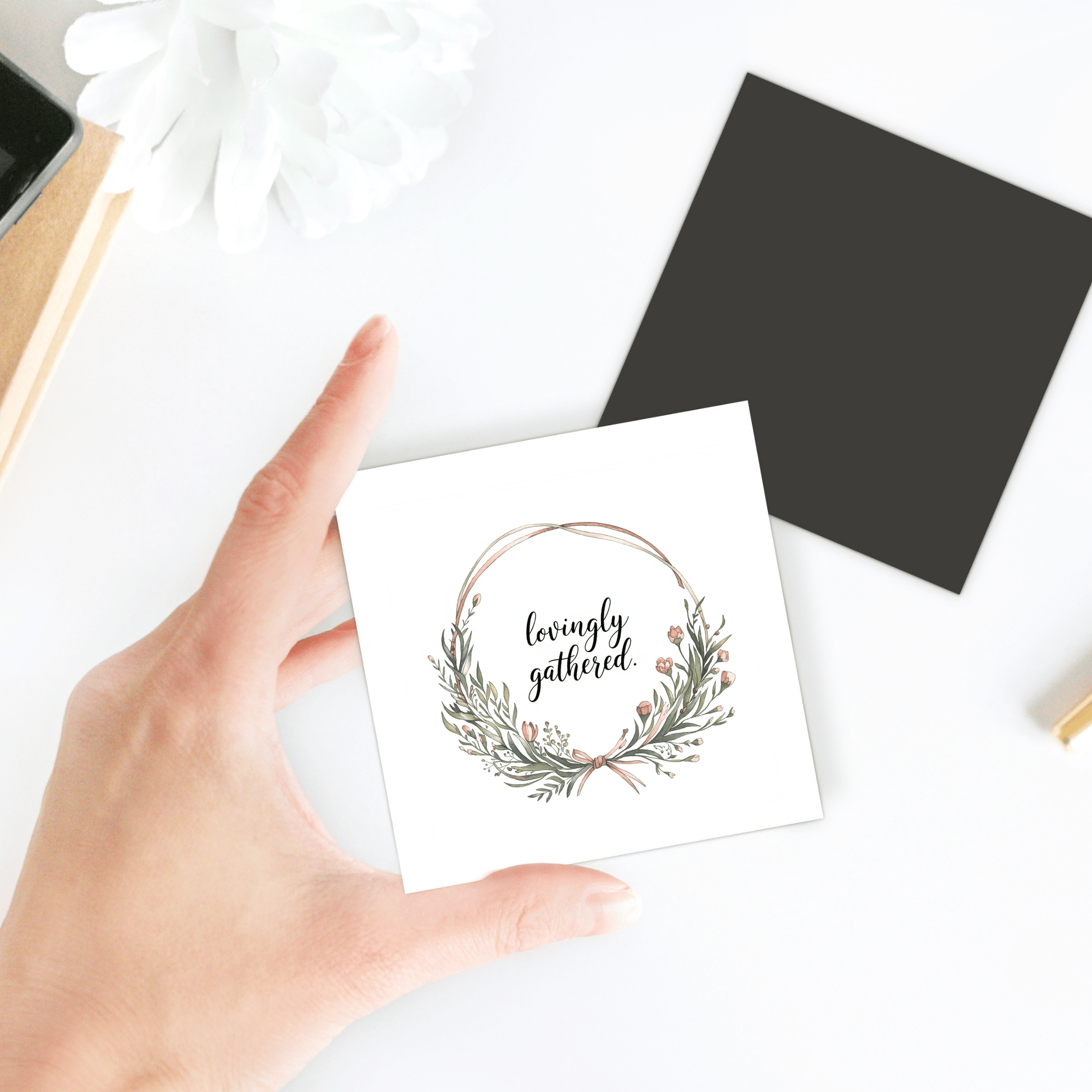A hand holds a white card featuring a wreath and “lovingly gathered” beside a Mirabilia Boutique Lovingly Gathered Magnet – Floral Wreath Quote for Fridge, a black envelope, and a white flower on a white surface.