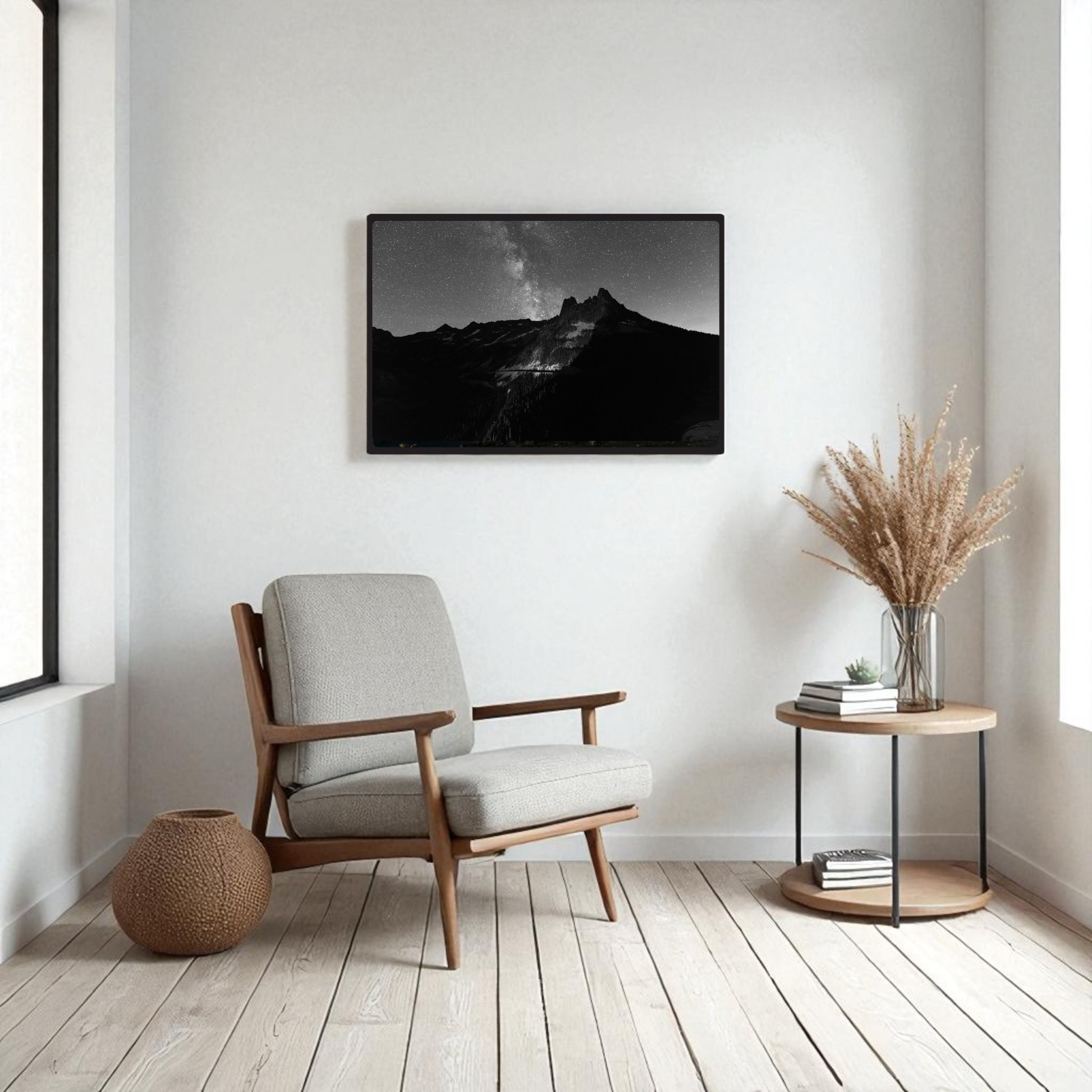 A minimalist living room features a cozy armchair, a round side table topped with a vase of dried grass, and a woven basket. On the wall hangs the striking “Liberty Bell Black & White Fine Art Print | Framed Art Print” by Mirabilia Boutique, capturing Liberty Bell Mountain in the North Cascades. Light wood floors and neutral decor complete the serene atmosphere.