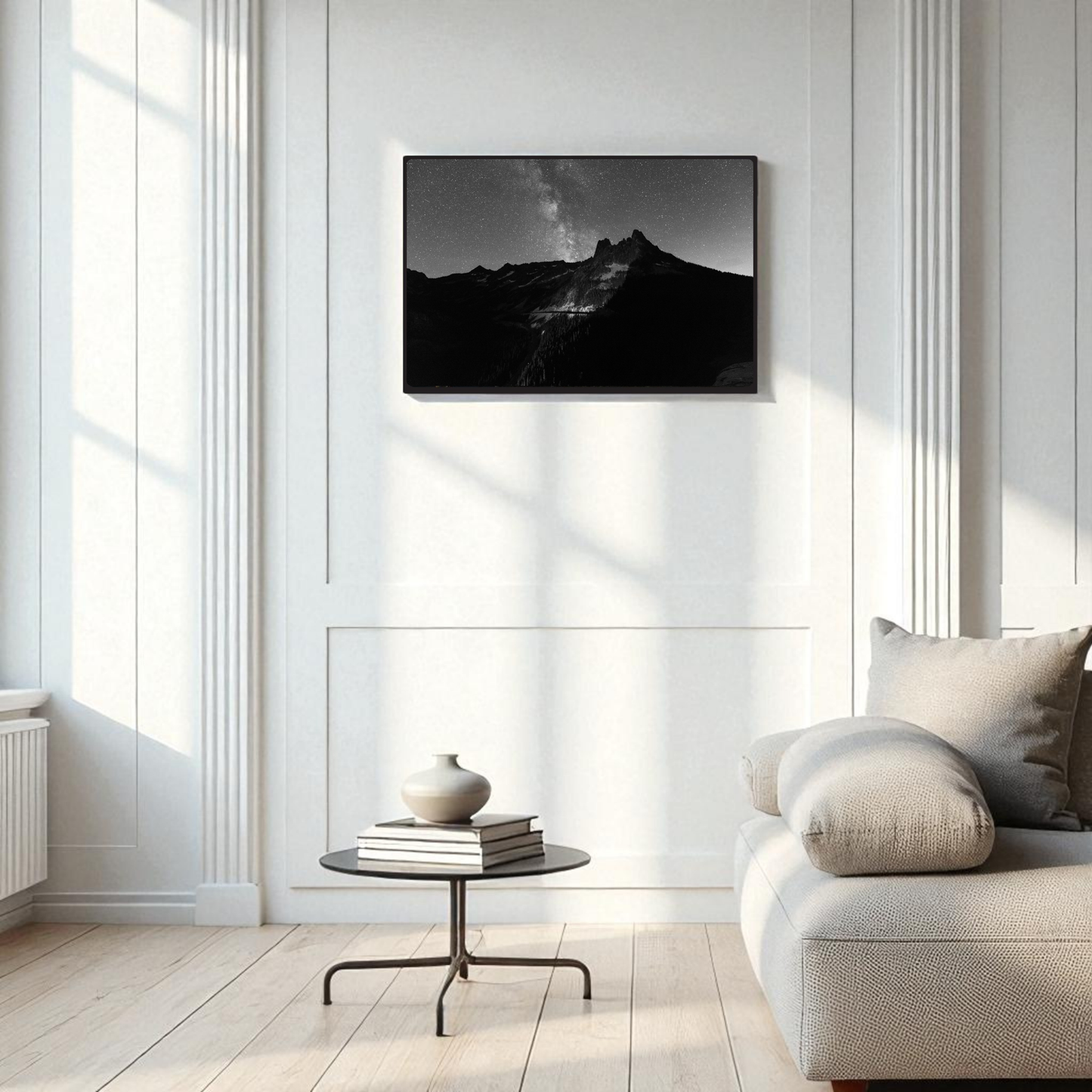 A modern living room features a light sofa, a round glass coffee table, and a vase, bathed in sunlight from large windows. Above the sofa, the Liberty Bell Black & White Fine Art Print by Mirabilia Boutique hangs on the white wall, its striking depiction of Liberty Bell Mountain in the North Cascades introducing natural elegance to the space.