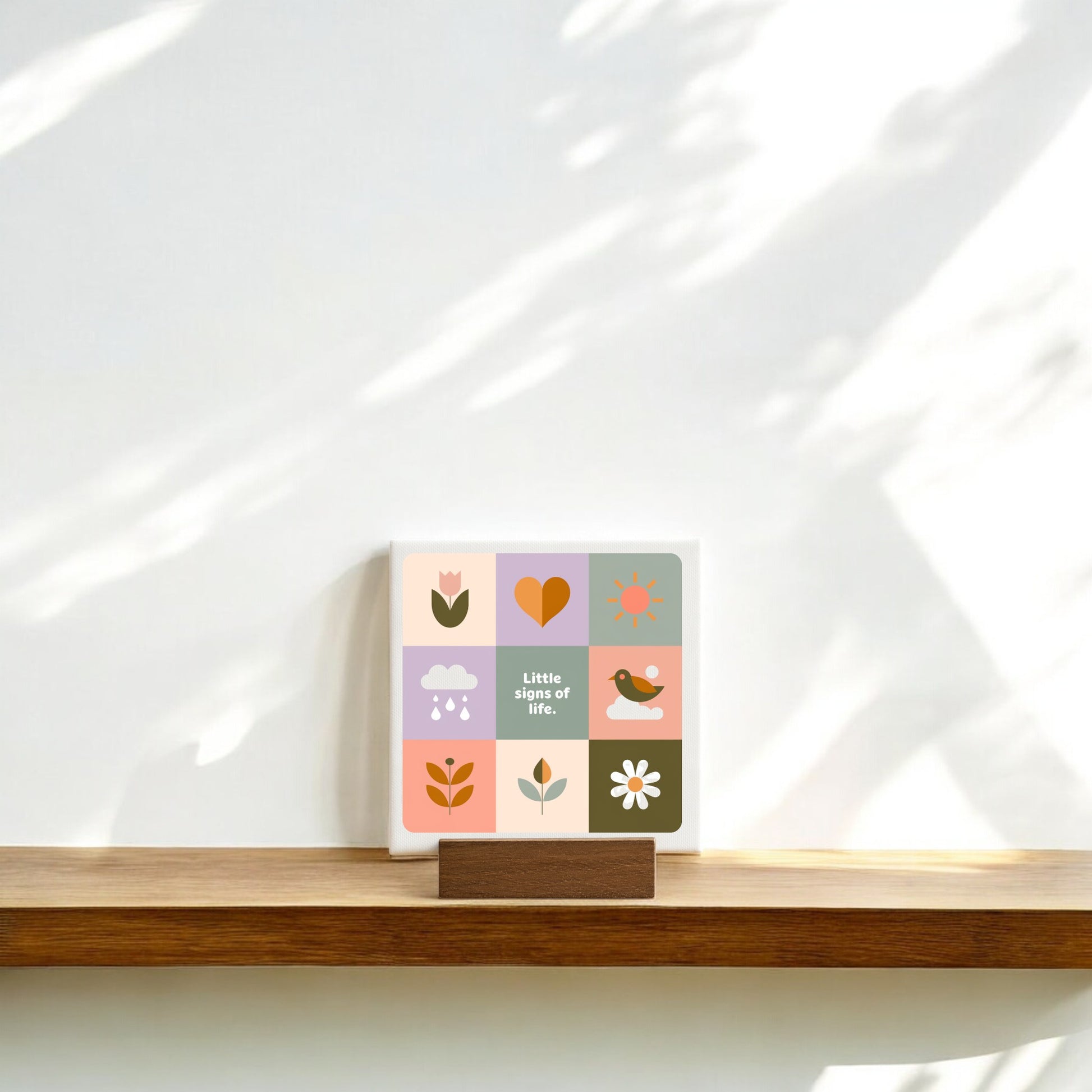 The Little Signs of Life Mini Canvas Sign with Base by Mirabilia Boutique features colorful illustrated squares and the phrase "Little signs of life." Perfect as inspirational desk decor, it brings modern charm to your home or office.