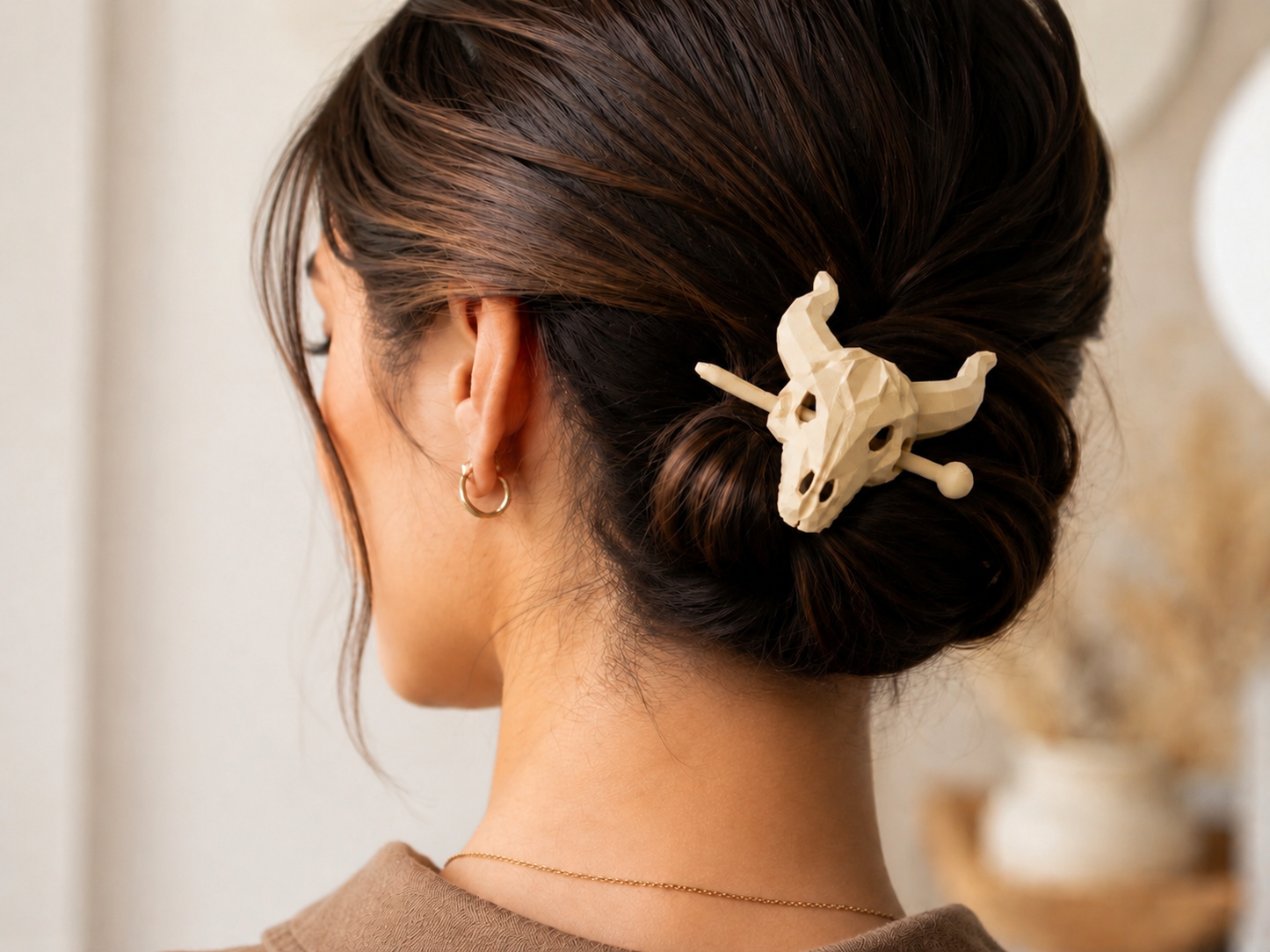 A woman with dark brown hair in a low bun, secured with the Mirabilia Boutique Longhorn Skull Hairpin | Western Theme Hair Accessory, wears a gold hoop earring and thin necklace, shown from behind.