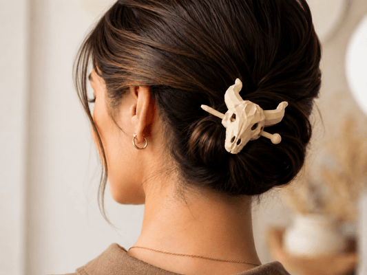 A woman with dark brown hair in a low bun, secured with the Mirabilia Boutique Longhorn Skull Hairpin | Western Theme Hair Accessory, wears a gold hoop earring and thin necklace, shown from behind.