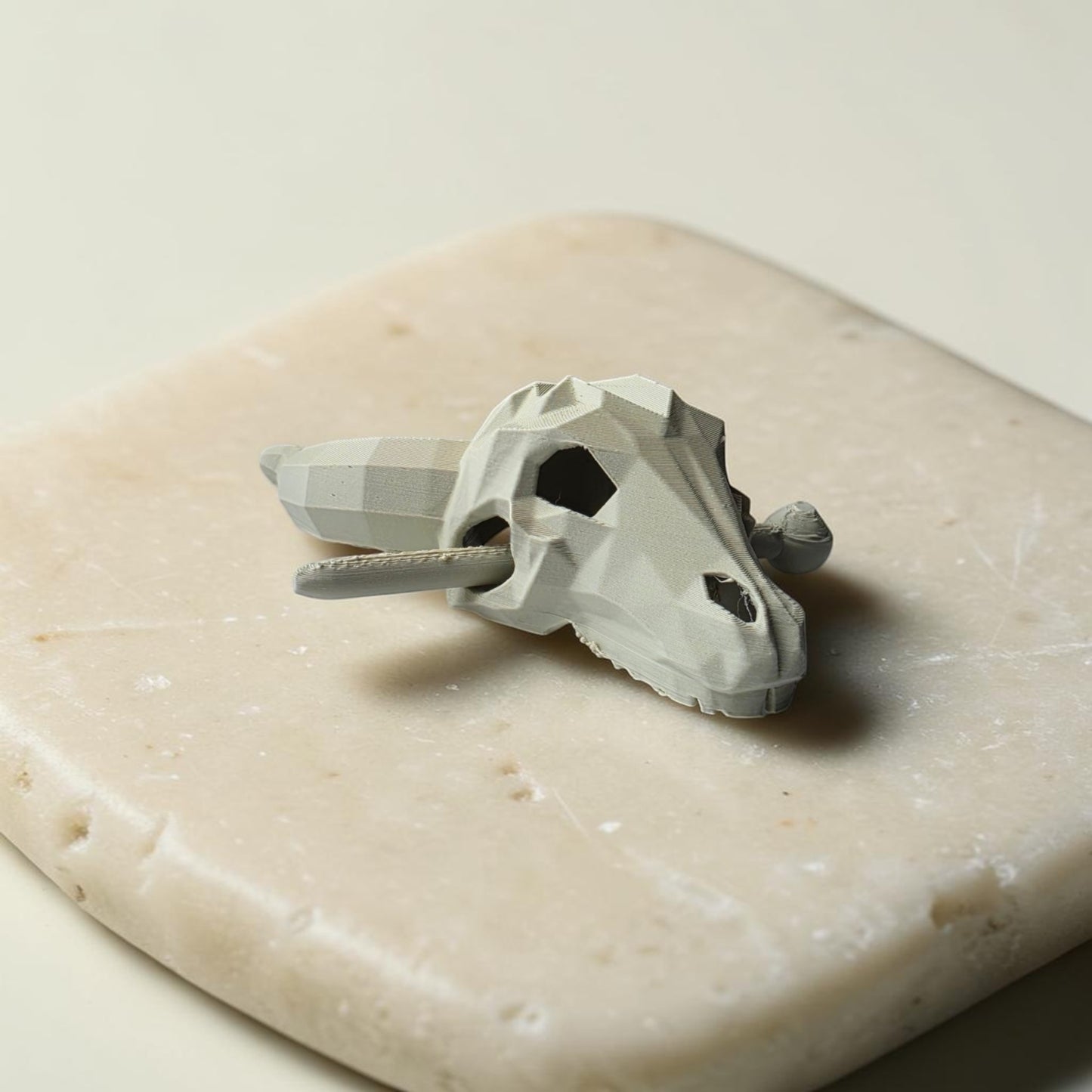 A small, geometric gray animal skull model with large eye sockets sits on a smooth beige stone, echoing the sculptural style of the Longhorn Skull Hairpin by Mirabilia Boutique—a striking Western-themed hair accessory.