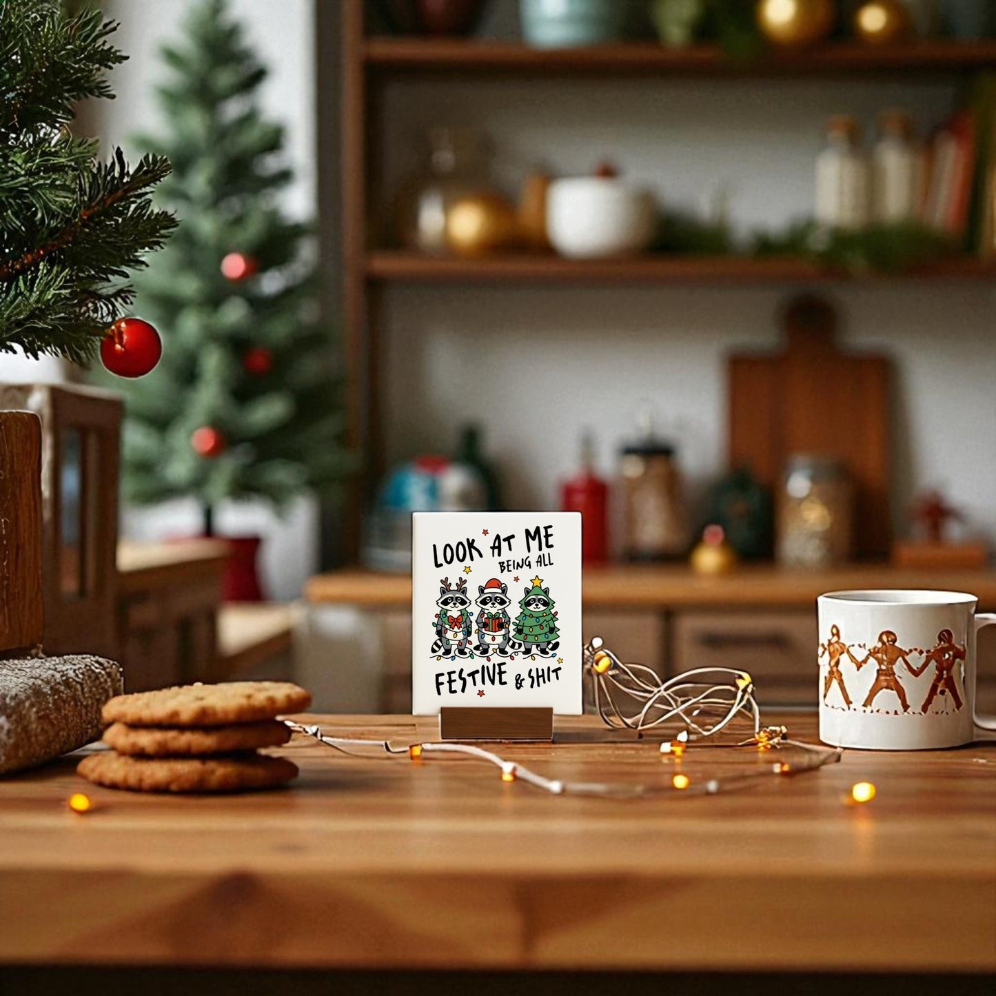 A cozy kitchen decorated for Christmas with a small tree, cookies, string lights, a Mirabilia Boutique “Look at Me Being Festive” funny raccoon sign, and a mug with gingerbread figures for extra cheer.