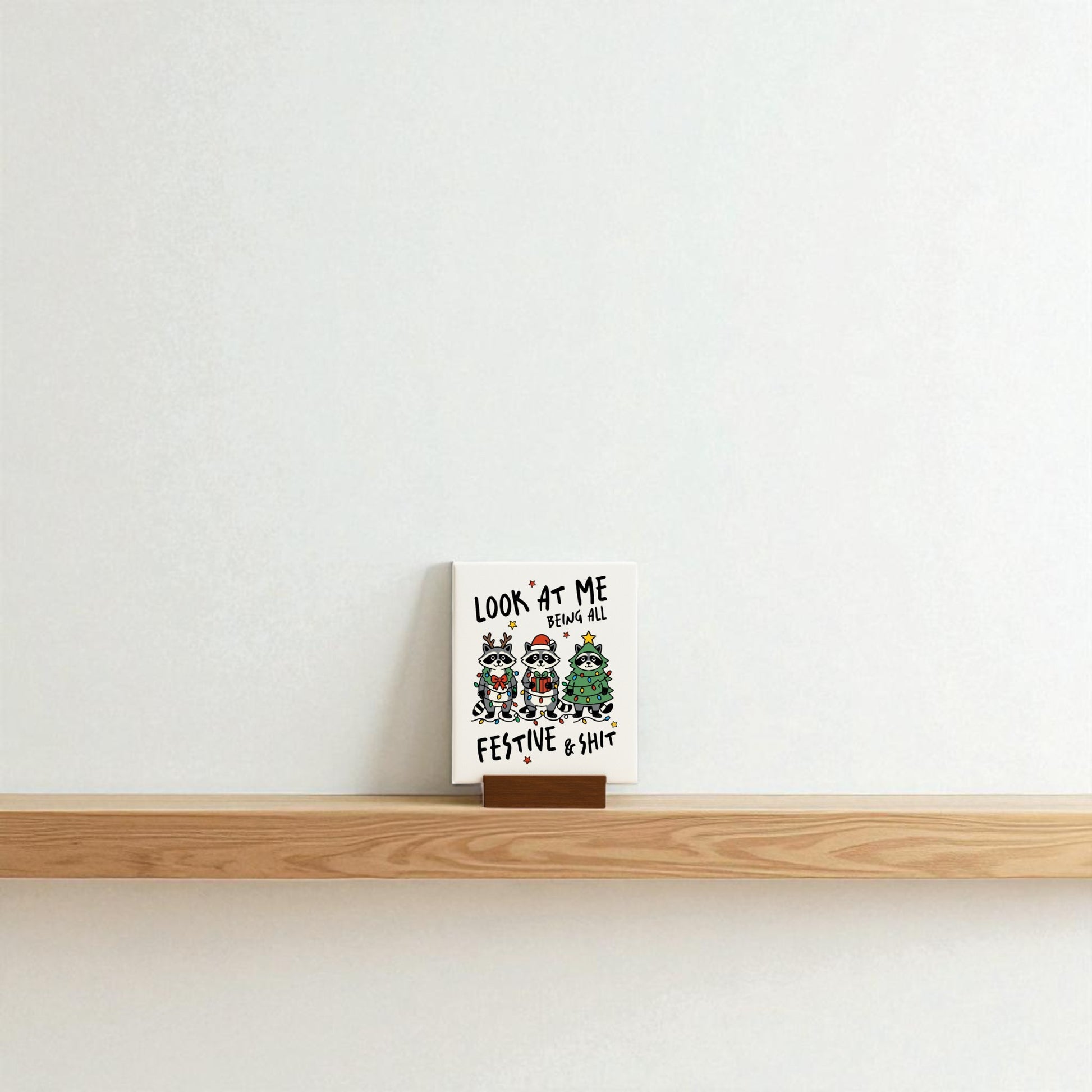 A Mirabilia Boutique "Look at Me Being Festive" Christmas raccoon sign sits on a wooden shelf against a plain white wall, making a snarky festive gift with cartoon animals in holiday attire and censored text.