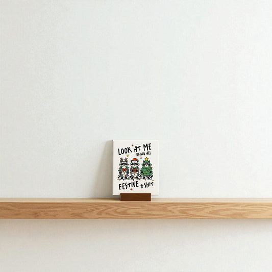 A Mirabilia Boutique "Look at Me Being Festive" Christmas raccoon sign sits on a wooden shelf against a plain white wall, making a snarky festive gift with cartoon animals in holiday attire and censored text.