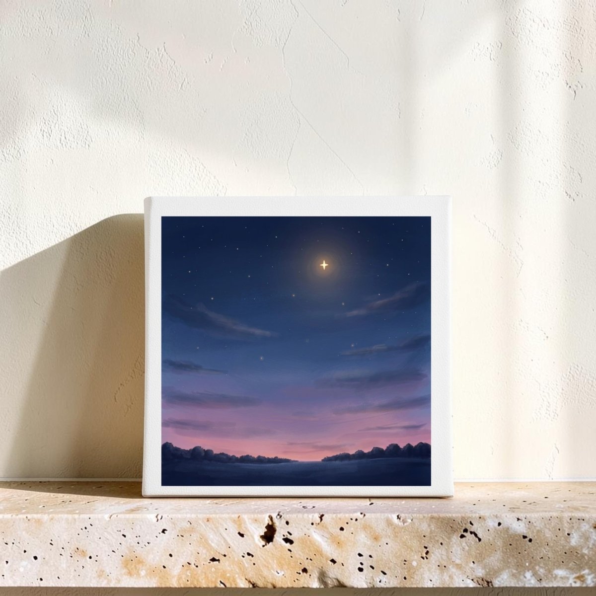 A square piece from Mirabilia Boutique, the “Look Up Celestial Canvas Wall Art,” rests on a stone shelf, showcasing a night sky with a luminous star above gentle clouds and a purple-pink horizon. Sunlight illuminates this tranquil artwork, creating soft shadows across the textured cream wall behind it.