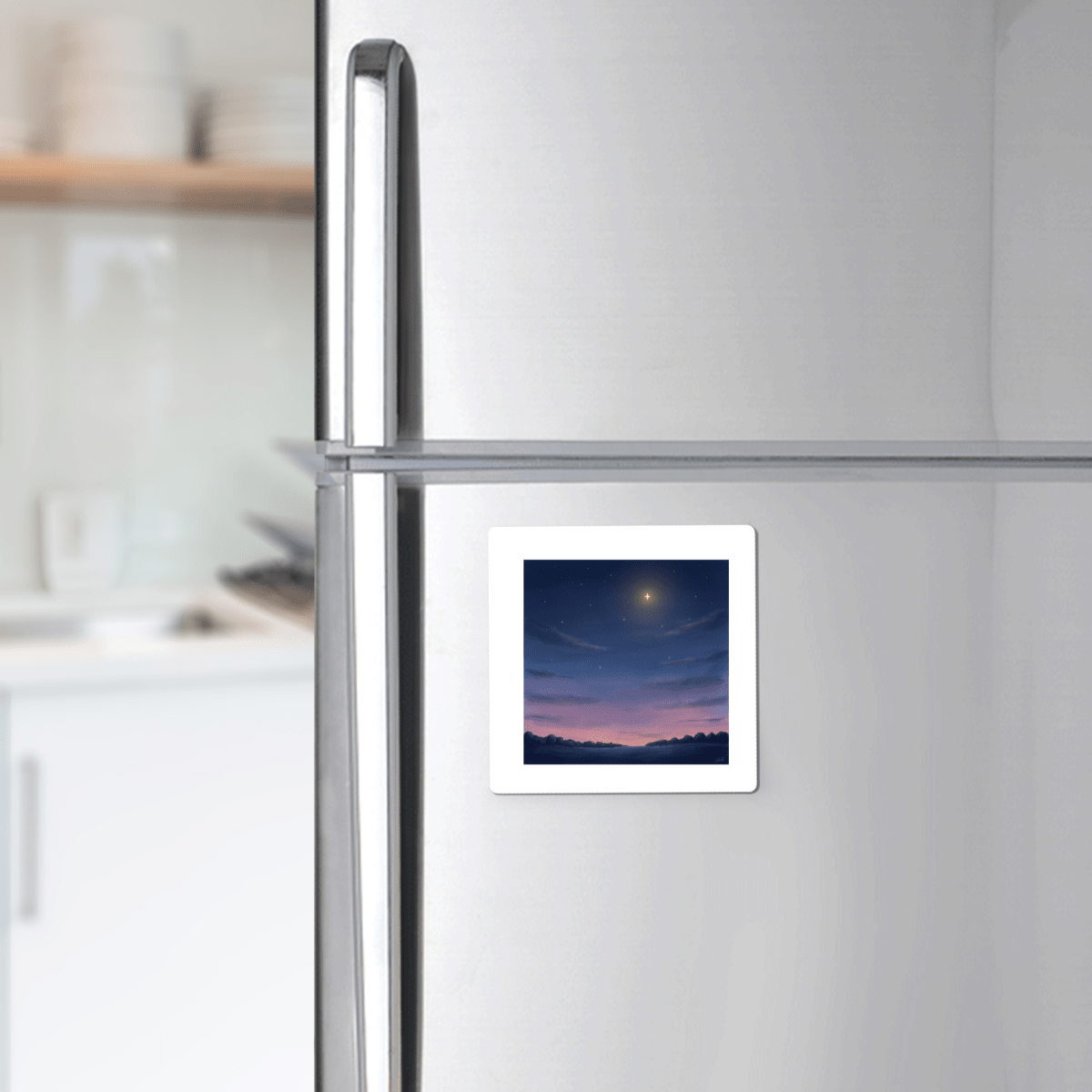 A silver refrigerator with a Mirabilia Boutique Look Up Celestial Magnet—a square twilight star design featuring a night sky scene above a dark landscape—displayed on the lower right door. Blurred white kitchen shelves can be seen in the background.