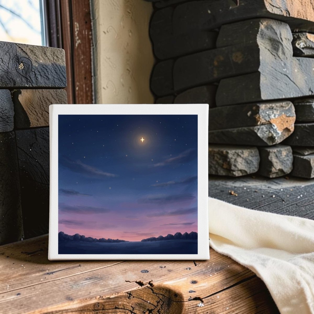 The Look Up Celestial Canvas Wall Art by Mirabilia Boutique—a small canvas featuring tranquil artwork of a glowing star in a twilight sky—rests on a rustic wooden table near stacked stones and a window.