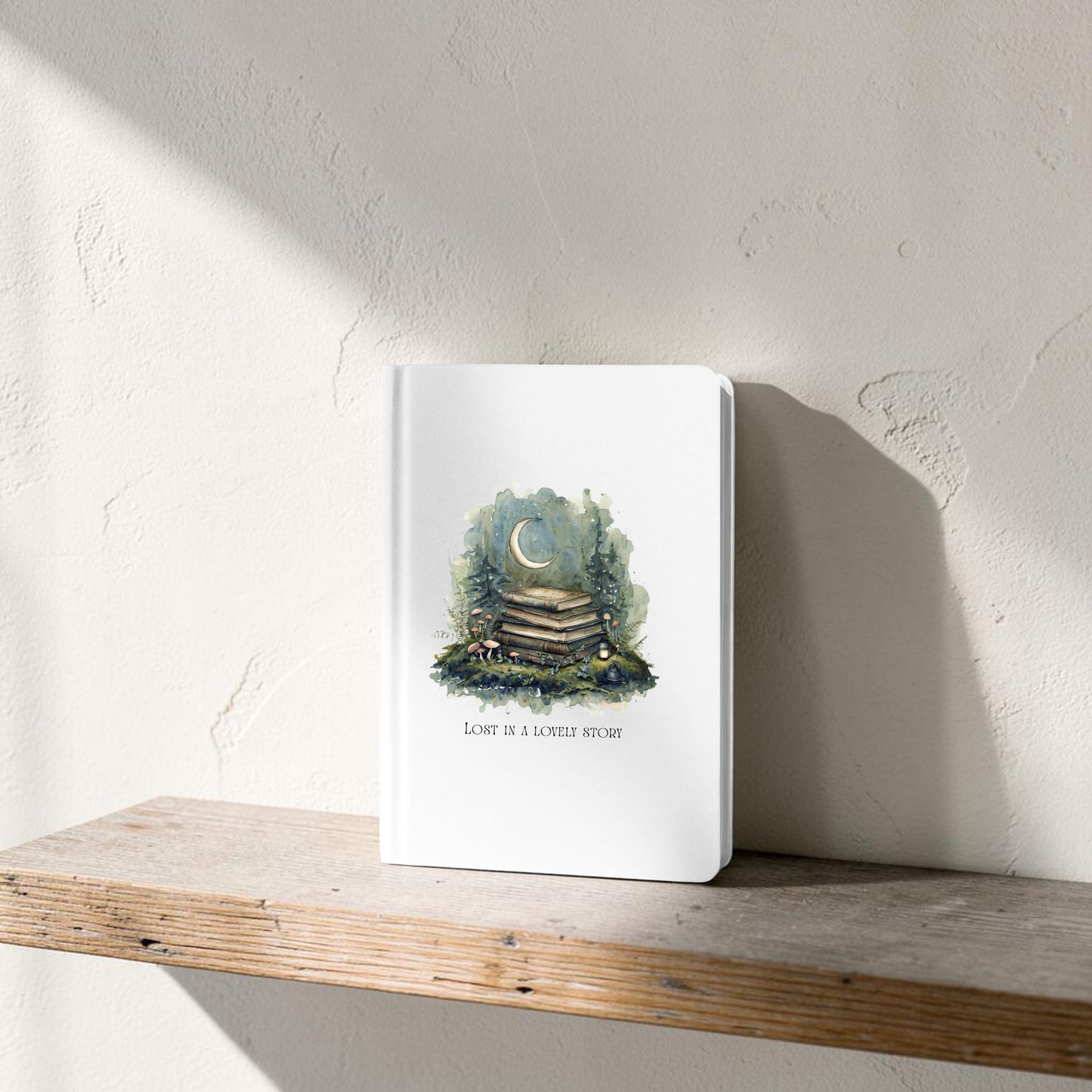 A Lost in a Lovely Story Journal by Mirabilia Boutique stands on a wooden shelf. Its white cover features an illustration of stacked books, greenery, and a crescent moon, with the words “Love is a longer story” displayed below the image.