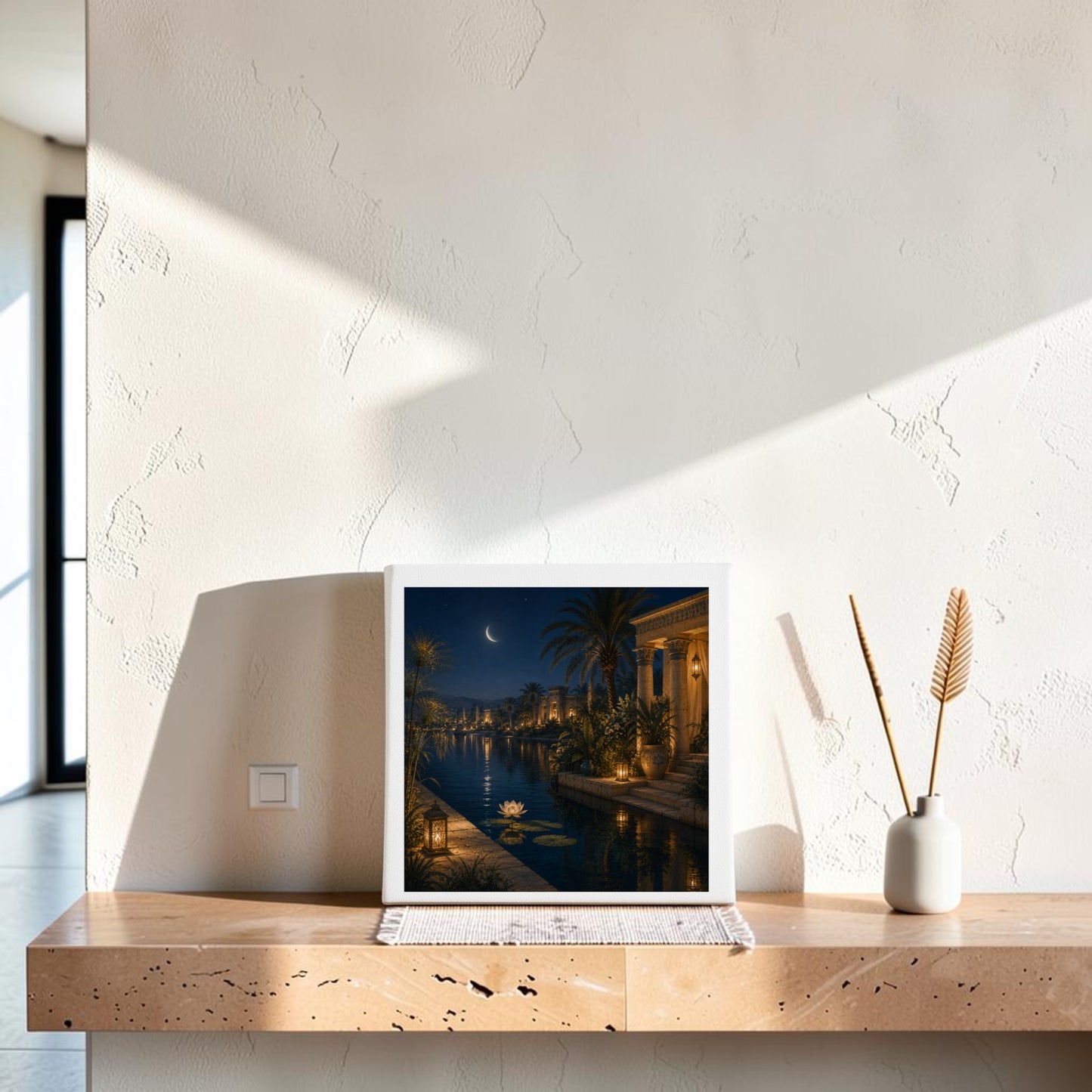 A framed print of “Lotus After Dark” from Mirabilia Boutique rests on a light wood shelf. Beside it, a beige vase holds two dried plants against a white textured wall, with sunlight casting gentle shadows—capturing the tranquil elegance of Egyptian canvas wall art.