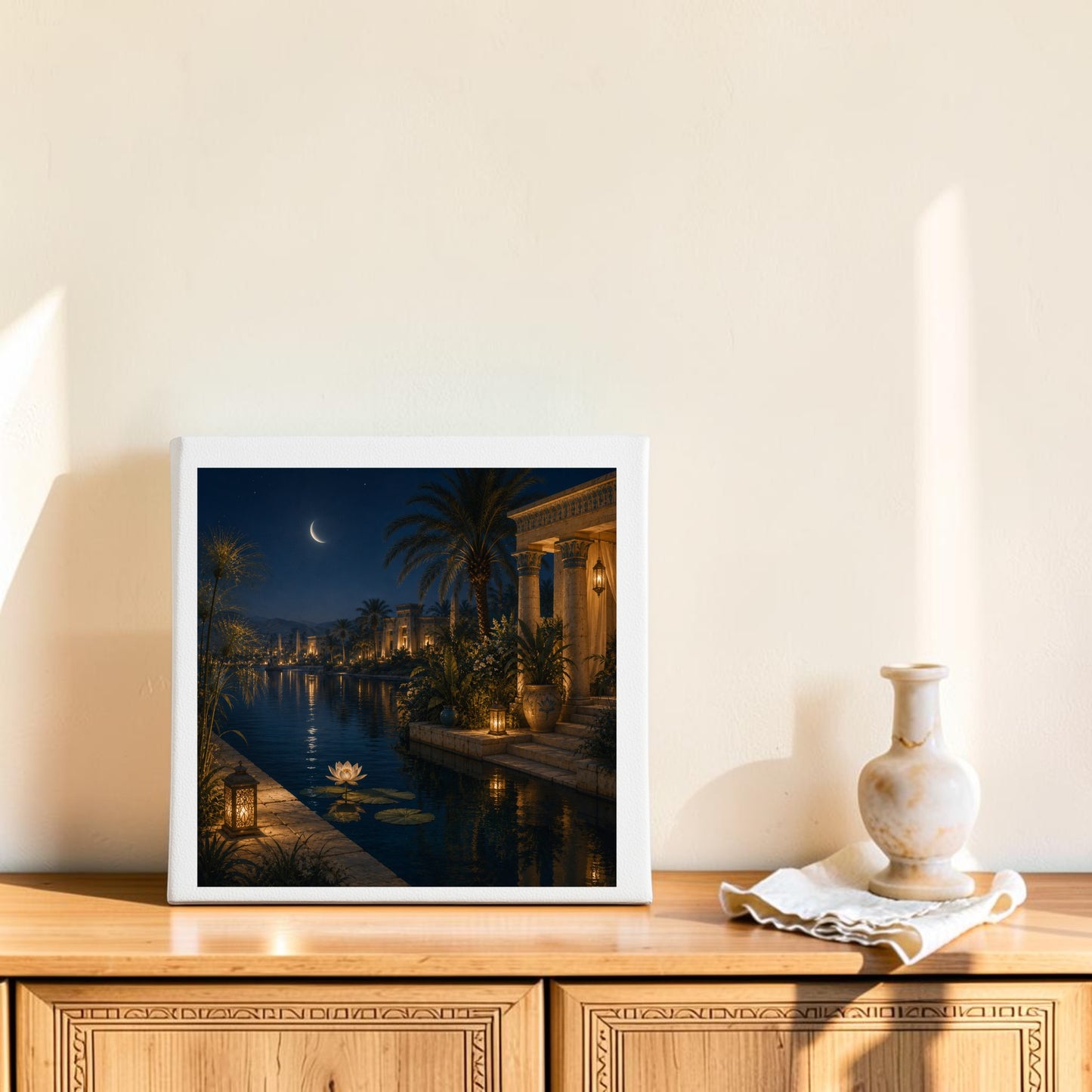 A square canvas titled “Lotus After Dark | Egyptian Canvas Wall Art” by Mirabilia Boutique—depicting a serene night scene with a crescent moon, palm trees, and tranquil waters reminiscent of the moonlit Nile—rests on a wooden sideboard alongside a ceramic vase and folded cloth, set against a cream-colored wall bathed in sunlight.