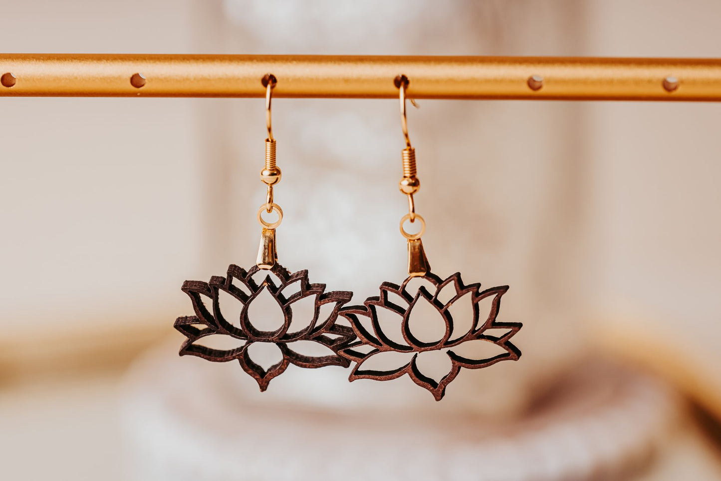 Two Laser Cut Lotus Flower Wood Earrings by Mirabilia Boutique, lightweight and boho, hang side by side from a golden rod. The blurred background highlights their delicate and intricate design.