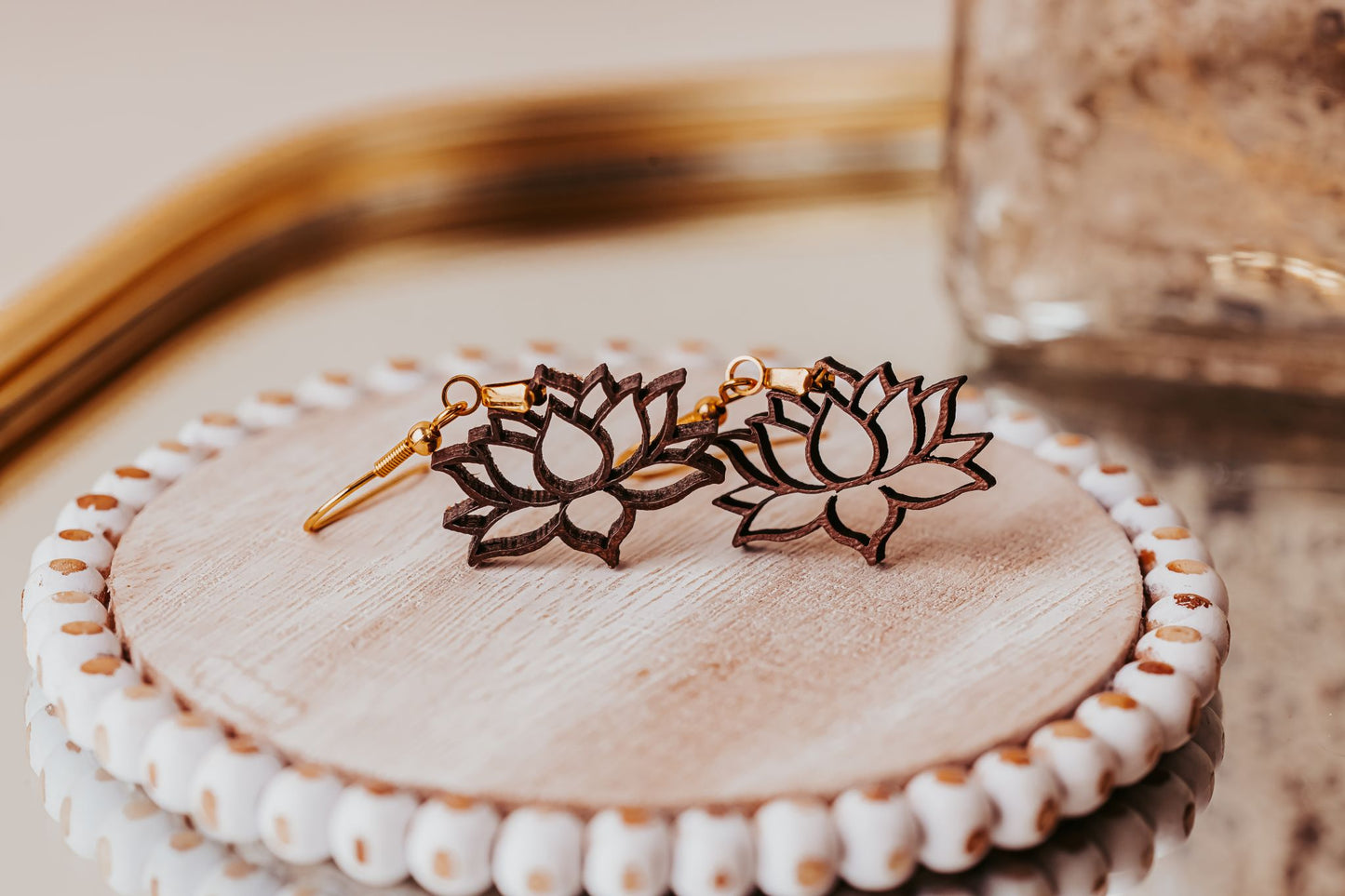 The Laser Cut Lotus Flower Wood Earrings by Mirabilia Boutique, lightweight and boho-style in gold and dark brown, rest on a round wood dish with a white beaded edge atop a reflective surface.
