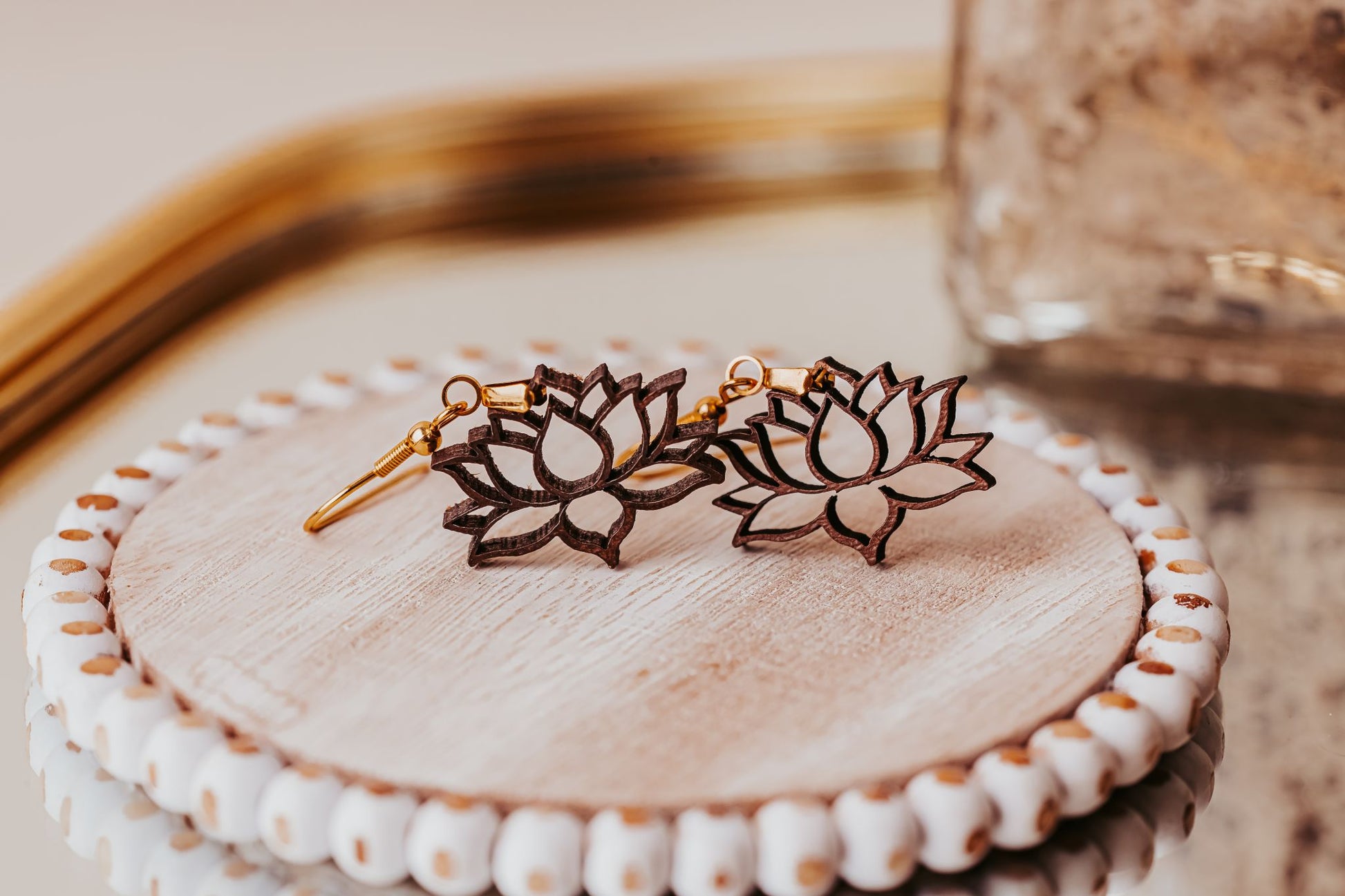 The Laser Cut Lotus Flower Wood Earrings by Mirabilia Boutique, lightweight and boho-style in gold and dark brown, rest on a round wood dish with a white beaded edge atop a reflective surface.