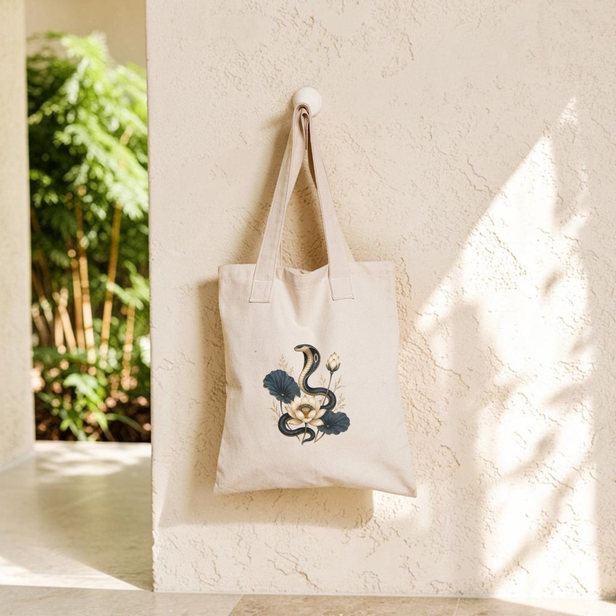 The Lotus Guardian Cobra Tote Bag by Mirabilia Boutique, featuring an ancient Egyptian snake and floral motif in beige, hangs on a wall hook as sunlight streams in and casts shadows across the textured wall.