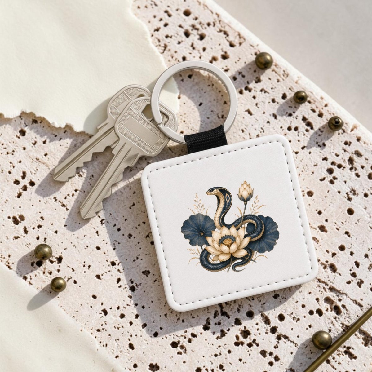 Two silver keys are attached to the Lotus Guardian Cobra Keychain by Mirabilia Boutique, which features a square white fob adorned with a black snake, cream flower design, and delicate lotus flowers from their Egyptian Lotus Art collection, all resting on a speckled beige surface.