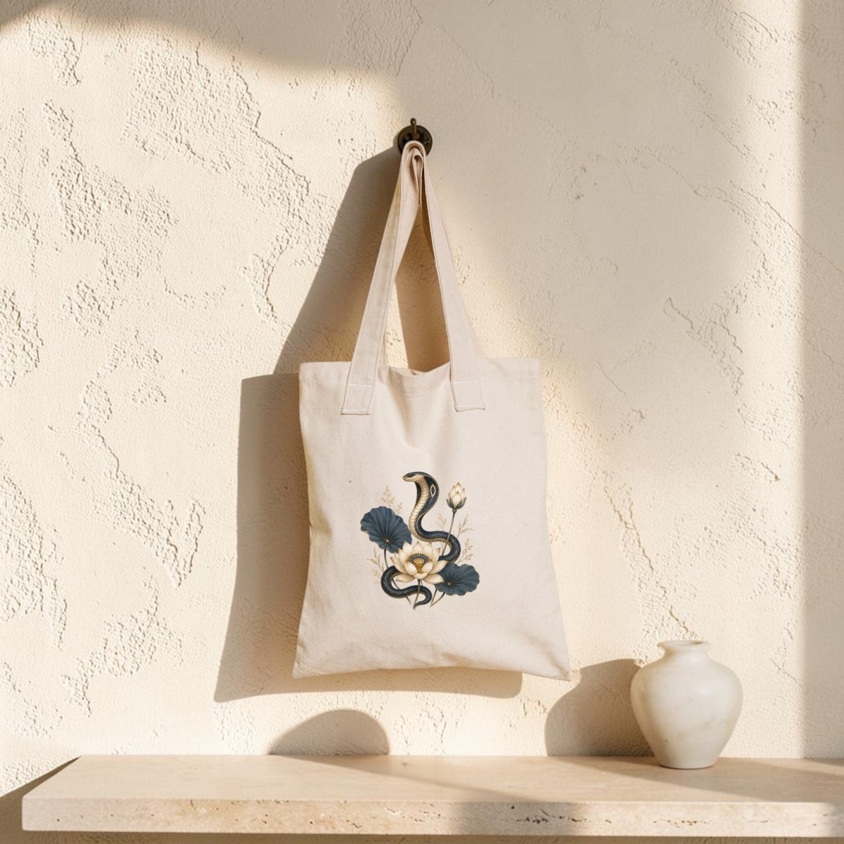 A Mirabilia Boutique Lotus Guardian Cobra Tote Bag, featuring an ancient Egyptian cobra and floral design in beige, hangs on a wall hook above a light stone shelf with a small white ceramic vase. Sunlight casts soft shadows across the textured wall.