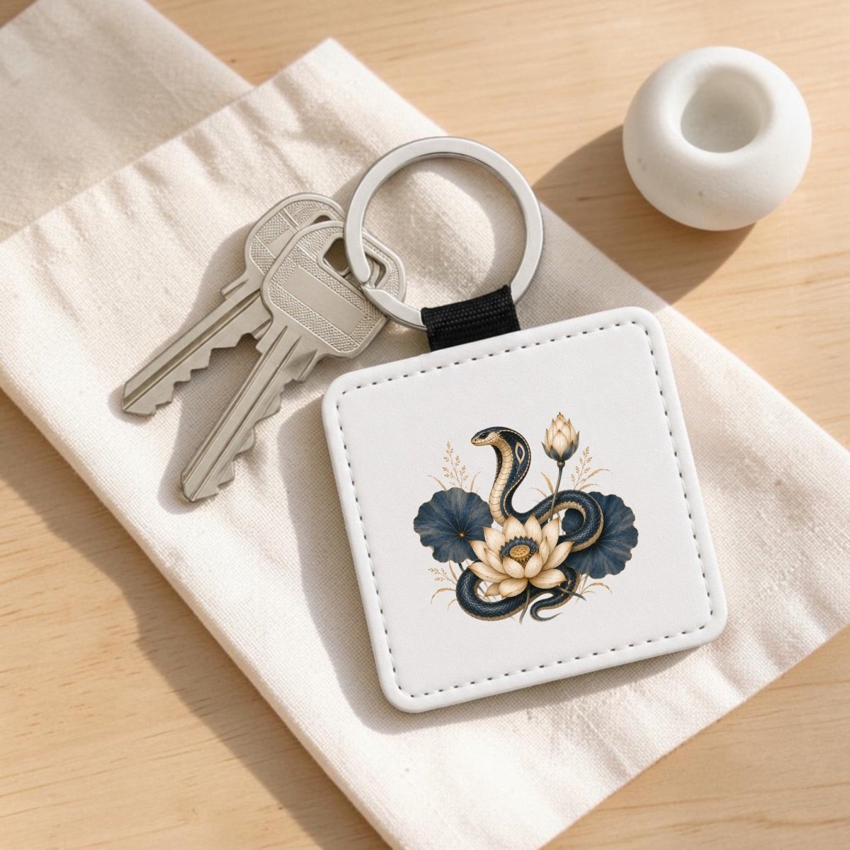 Restyled Sentence: The Lotus Guardian Cobra Keychain from Mirabilia Boutique rests on a white cloth, showcasing its striking Egyptian lotus art design— a black cobra entwined with cream and blue floral motifs accented with lapis lazuli hues.