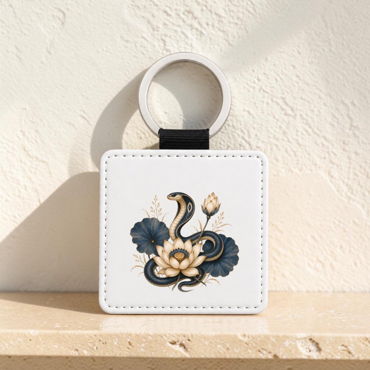 The Lotus Guardian Cobra Keychain | Egyptian Lotus Art by Mirabilia Boutique is a square white keychain displaying a striking black cobra coiled among delicate white lotus flowers and vibrant lapis lazuli-blue leaves. It is elegantly positioned on a beige surface against a light-colored wall.
