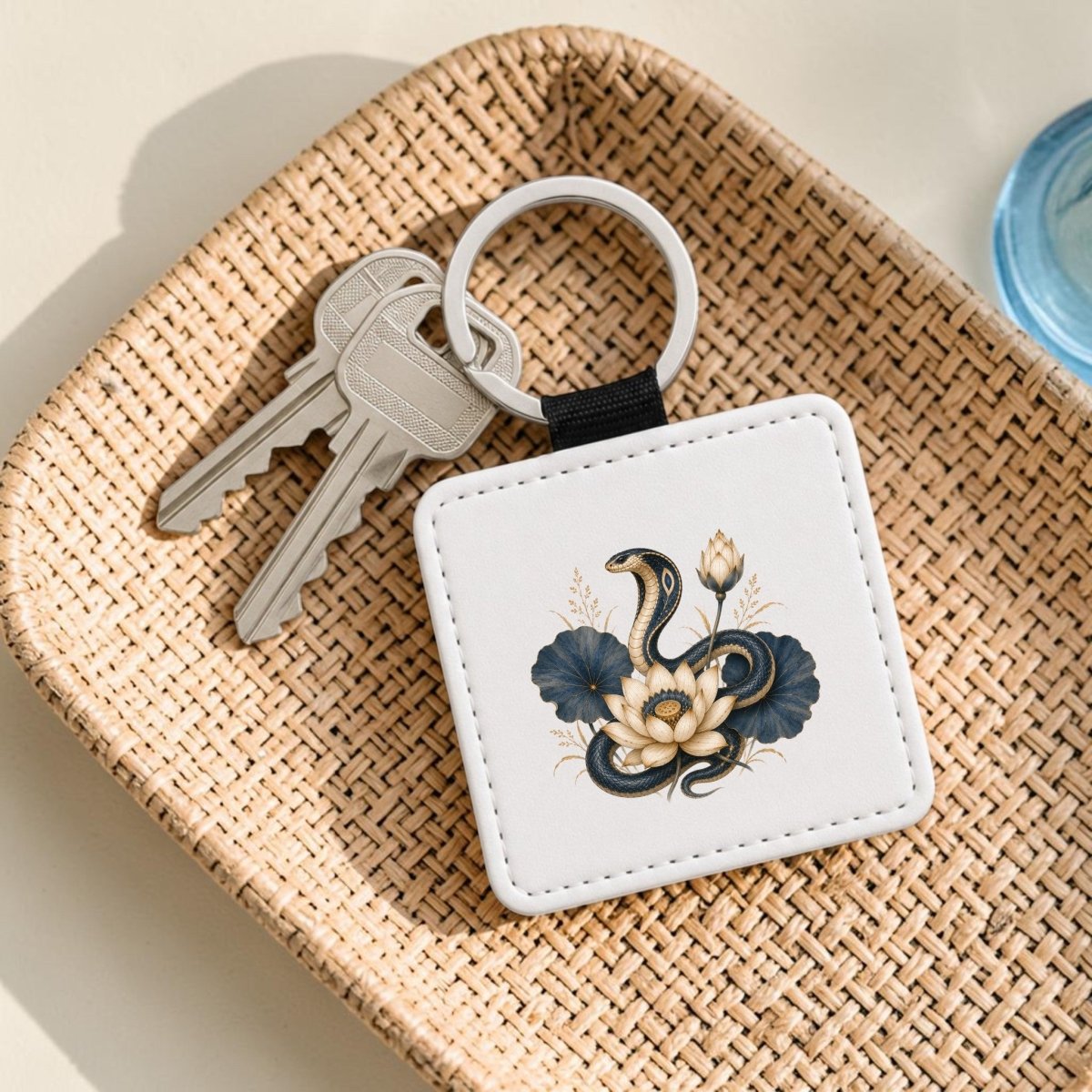 A square keychain featuring a snake and lotus flower design from Mirabilia Boutique’s Lotus Guardian Cobra Keychain | Egyptian Lotus Art rests in a woven tray beside two silver keys.
