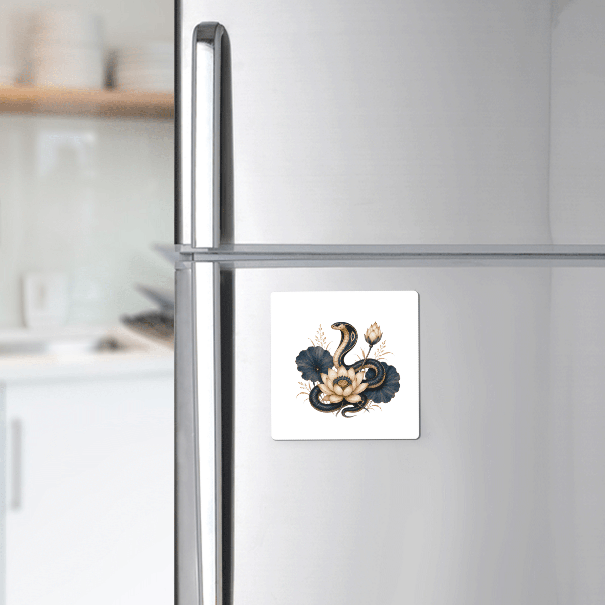 A silver refrigerator displays the Mirabilia Boutique Lotus Guardian Cobra Magnet, a square magnet featuring a black and white floral and snake design inspired by Egyptian lotus art. This accent is perfect for adding a touch of Lapis & Lotus-inspired décor. Kitchen shelves with dishes appear blurred in the background.
