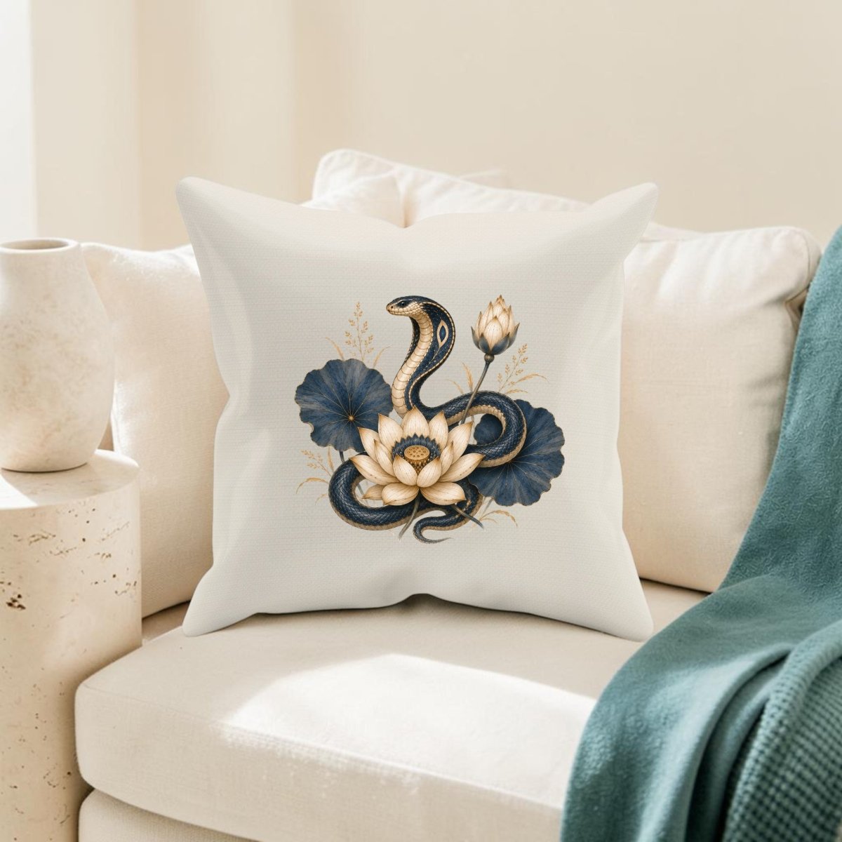 A white couch displays the Lotus Guardian Cobra Pillow | Ancient Egyptian Art from Mirabilia Boutique, which features a coiled snake among a white lotus flower and dark blue leaves, channeling the elegance of ancient Egyptian decor; a vase and green throw blanket complete the scene.