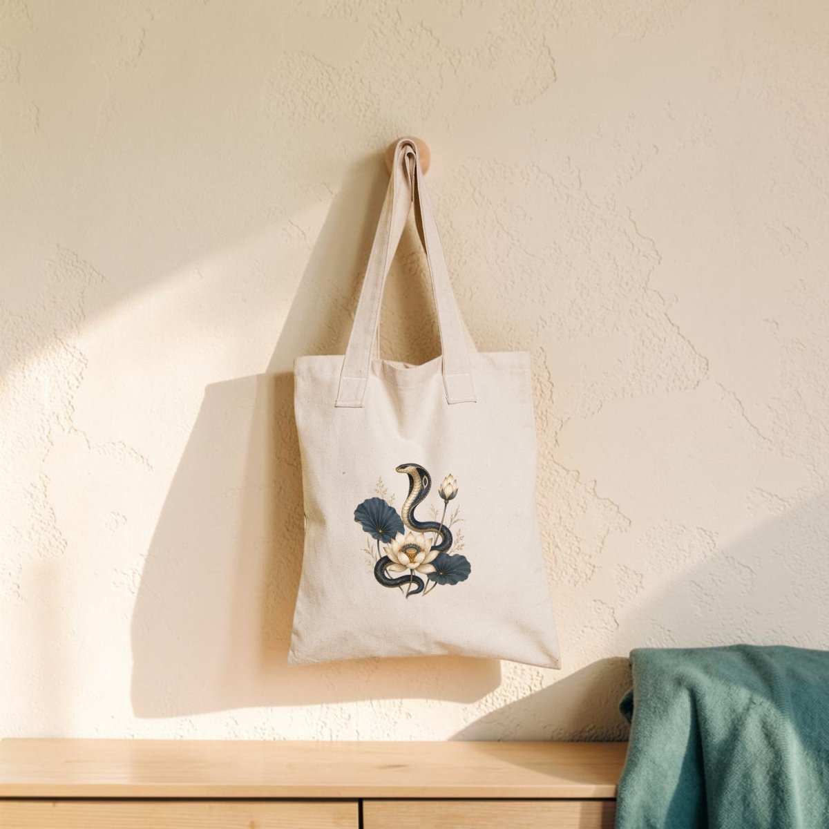 A Lotus Guardian Cobra Tote Bag from Mirabilia Boutique, featuring an ancient Egyptian snake and floral design in beige, hangs on a textured wall above a light wooden surface draped with a green cloth. Sunlight casts soft shadows across the scene.