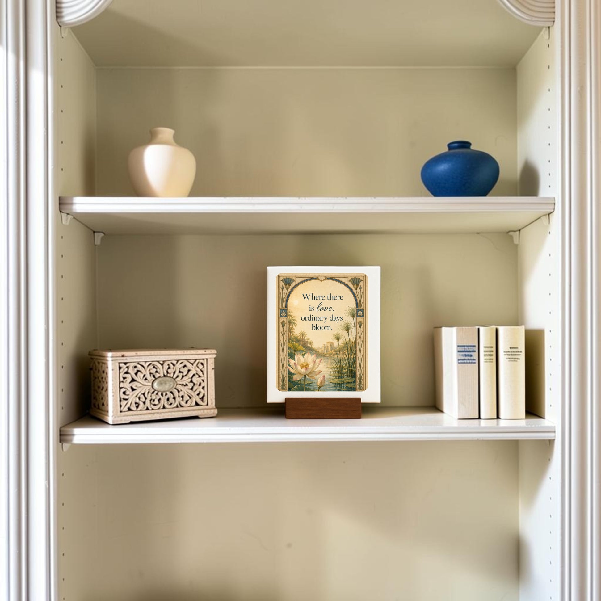 A decorative shelf featuring the Mirabilia Boutique Where There Is Love | Lotus Mini Sign from the Lapis & Lotus Collection, displayed alongside a cream vase, a blue vase, a carved box, and several books. The framed quote reads, “Where there is fear, ordinary days bloom.”.
