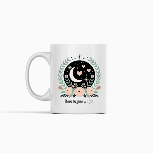 The Love Begins Within Mug from Mirabilia Boutique features a crescent moon, hearts, stars, and pink flowers with green leaves, plus the quote "Love begins within." A perfect self-love gift for any occasion. Shown on a white background.