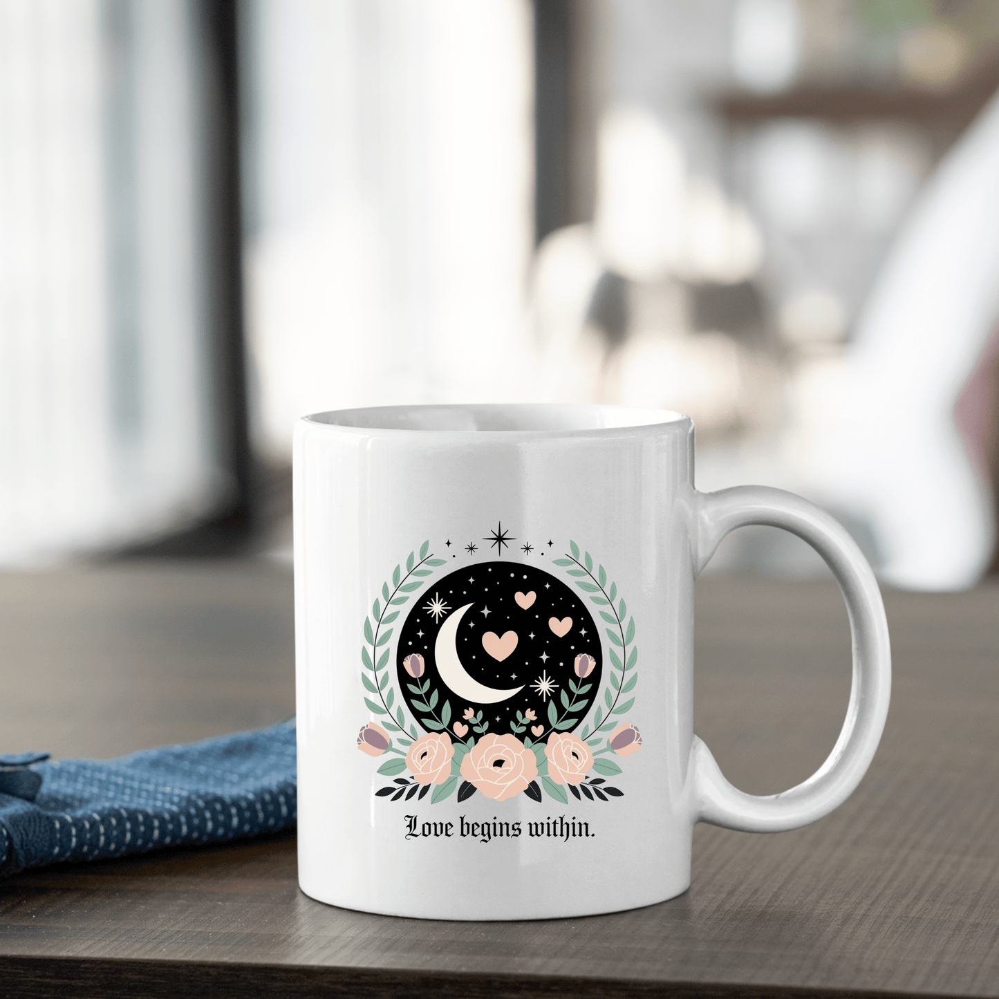 The Love Begins Within Mug by Mirabilia Boutique sits on a table, adorned with a crescent moon, hearts, stars, and pink flowers plus “Love begins within.” This self-love gift is perfect for mindfulness. A blue cloth is placed beside the mug.