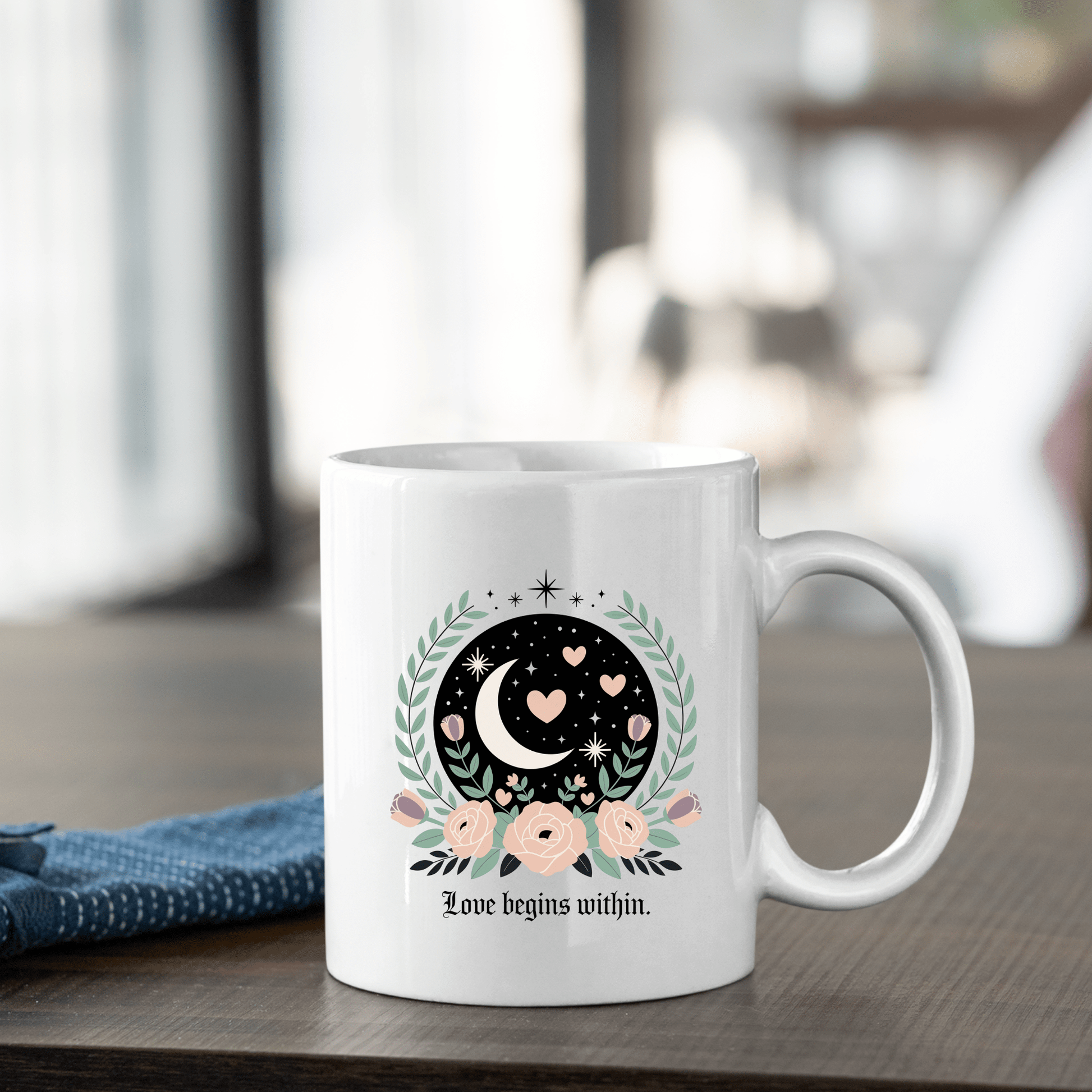 The Love Begins Within Mug by Mirabilia Boutique sits on a table, adorned with a crescent moon, hearts, stars, and pink flowers plus “Love begins within.” This self-love gift is perfect for mindfulness. A blue cloth is placed beside the mug.