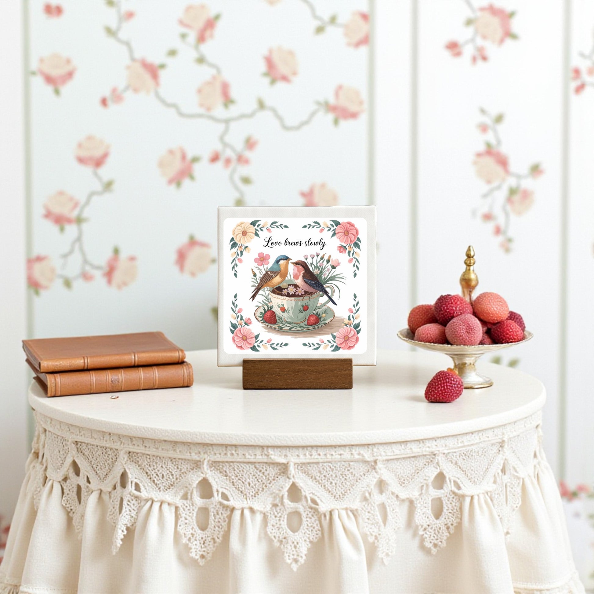 A round table with a lace tablecloth displays two stacked books, Mirabilia Boutique’s “Love Brews Slowly Mini Canvas” (Romantic Teacup Bird Art 4x4), a dish of pink sweets, and is set against floral wallpaper with pink roses.